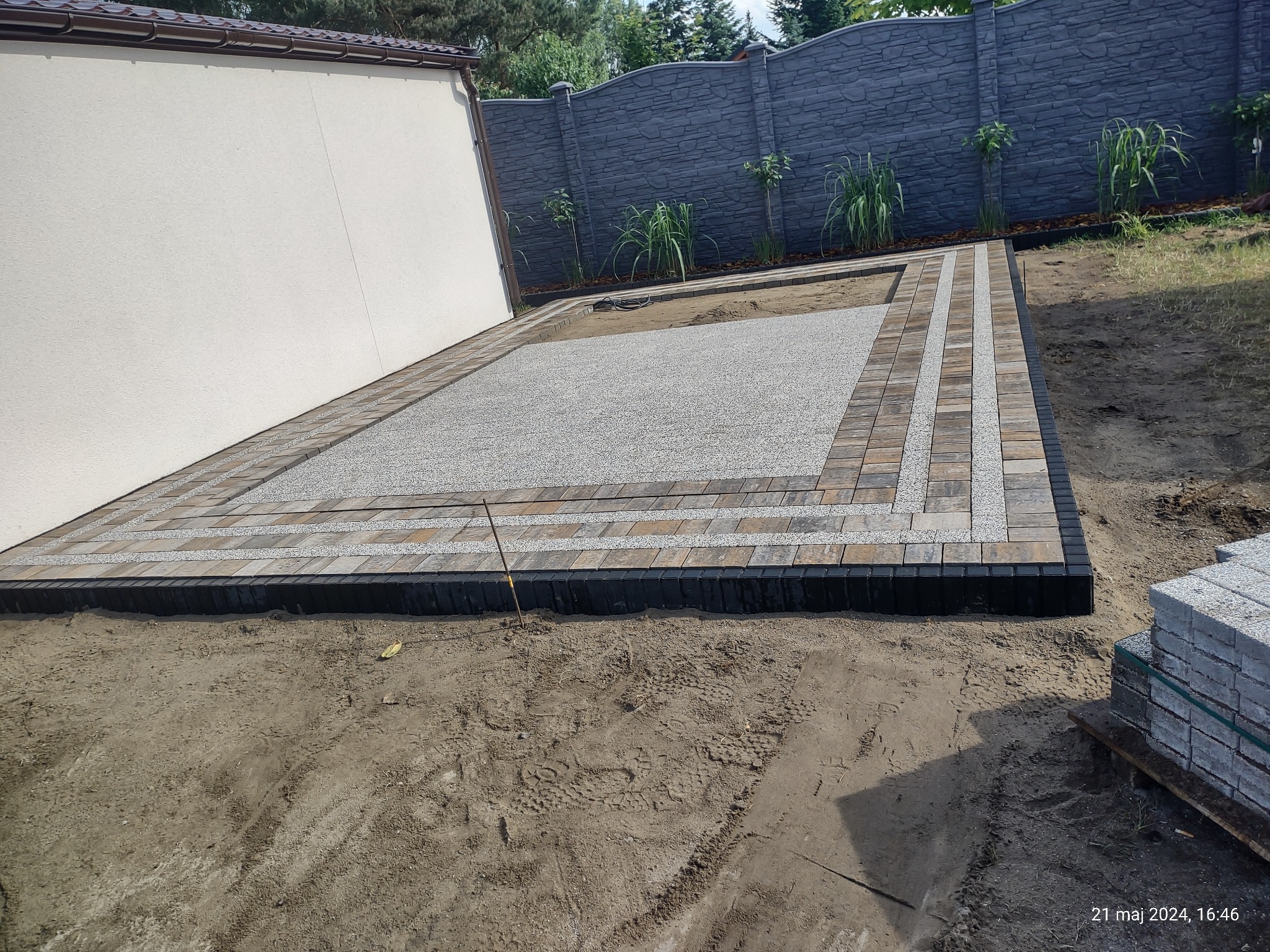 Fragment podwórka z wykończoną obwódką z kostki brukowej i wysypanym drobnym kamieniem, widoczne zapasy kostki i świeżo posadzone rośliny wzdłuż ogrodzenia.