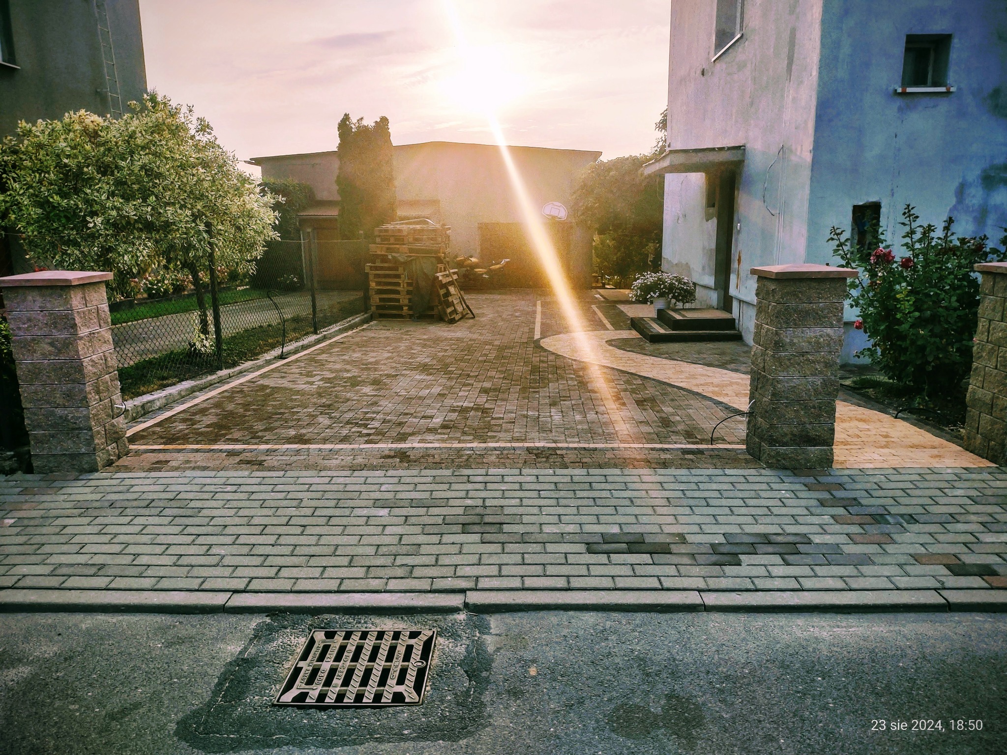 Nowo ułożona szara kostka brukowa prowadząca do domu z elementami jasnej kostki, widoczna studzienka kanalizacyjna, słoneczna poświata w tle.