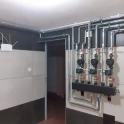 Rozdzielacz z trzema pompami obiegowymi zamontowany na ścianie w pomieszczeniu z instalacją rurową pod sufitem, widoczny router Wi-Fi.