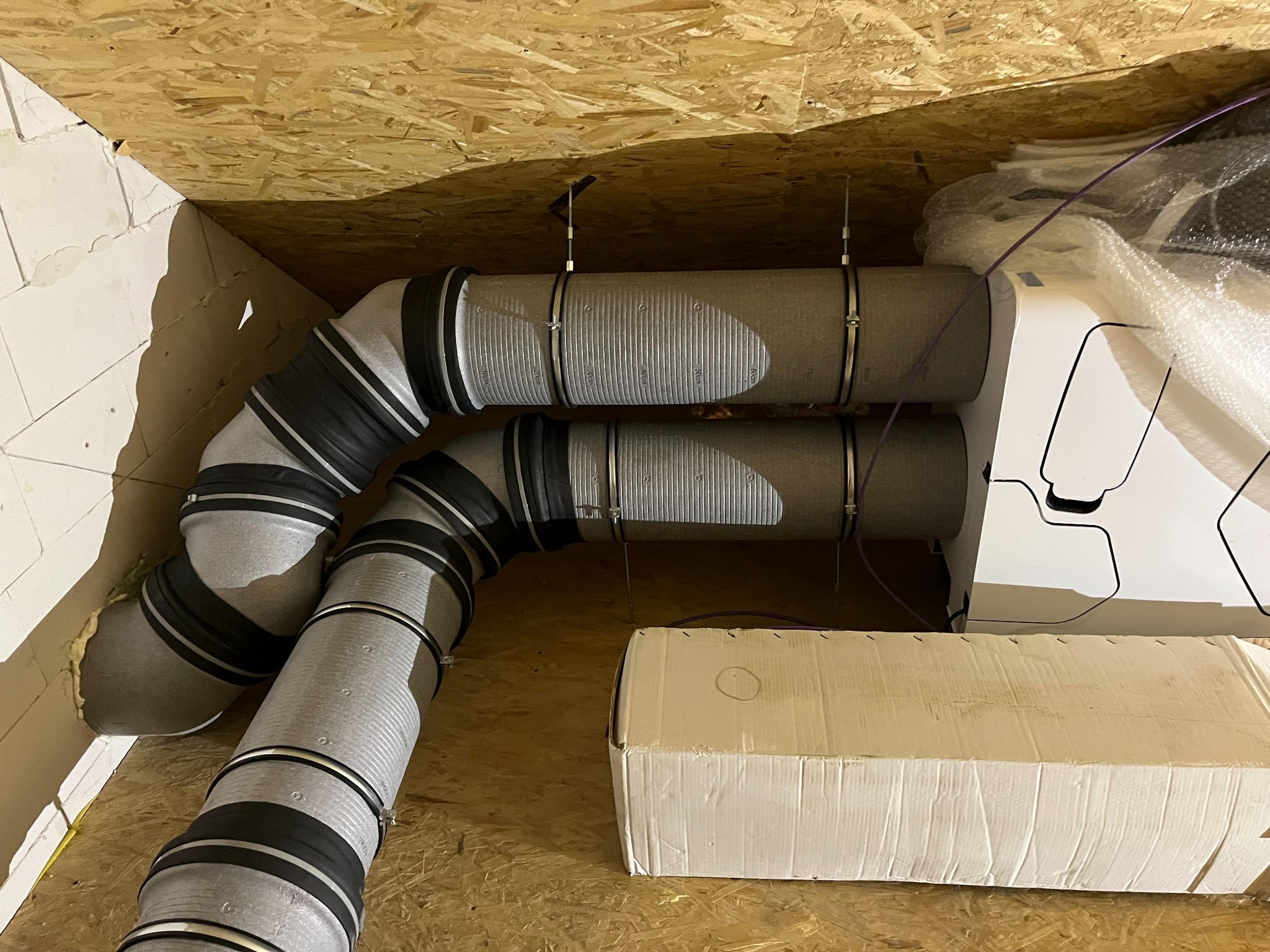Szare, elastyczne rury wentylacyjne z czarnymi wzmocnieniami, zamontowane pod drewnianym stropem, z widocznym białym urządzeniem i kartonem. Instalacja w trakcie montażu.