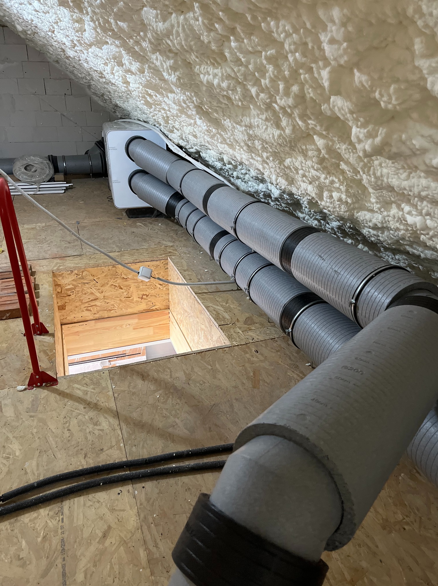 Rekuperacja na poddaszu: biała centrala wentylacyjna, szare rury wentylacyjne z czarnymi obejmami, izolacja piankowa na skosach dachu, właz na poddasze.