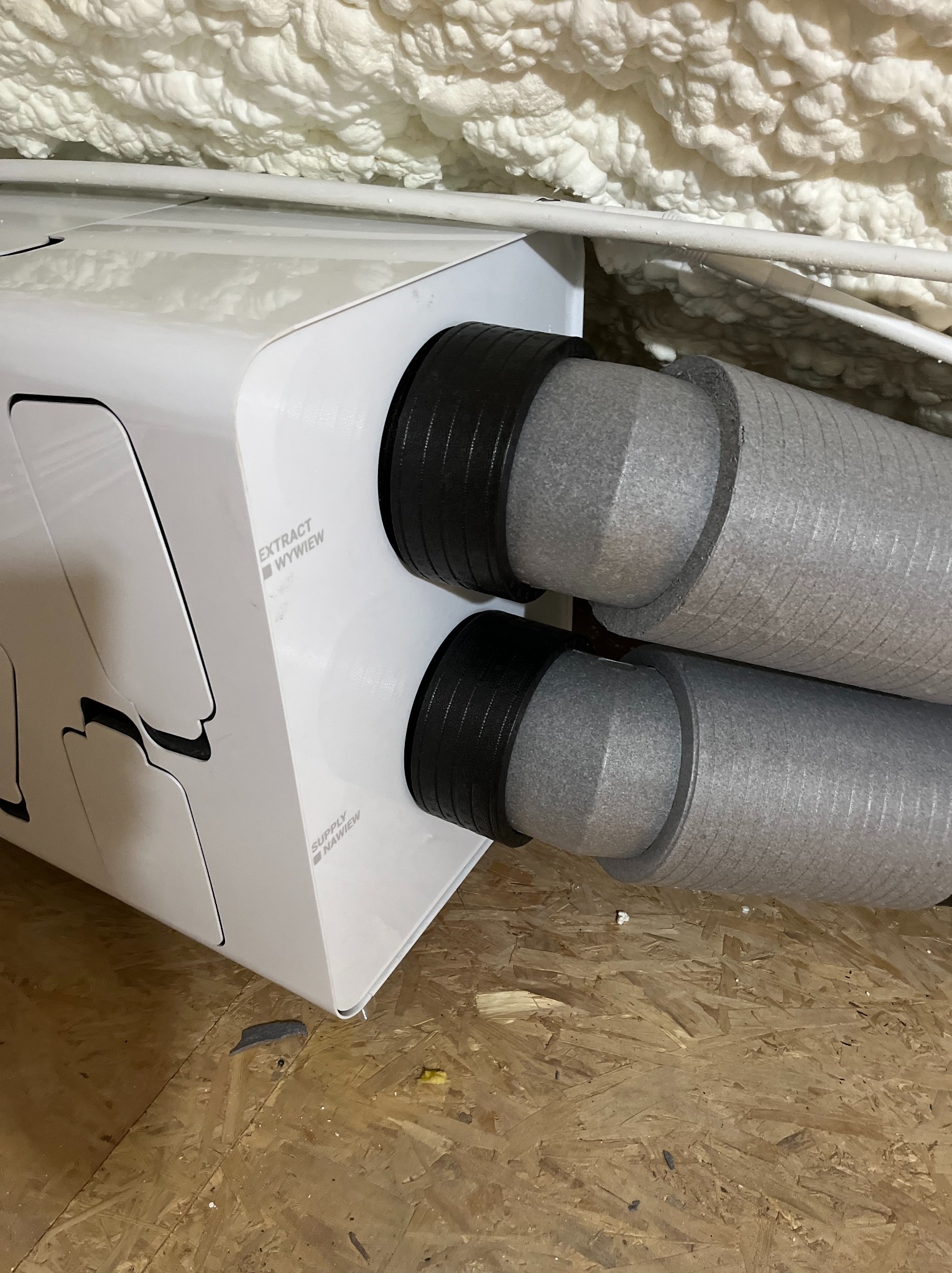 Biała jednostka rekuperacji z podłączonymi przewodami wentylacyjnymi, zamontowana pod stropem ocieplonym pianką PUR, widok z dołu na drewnianą podłogę.