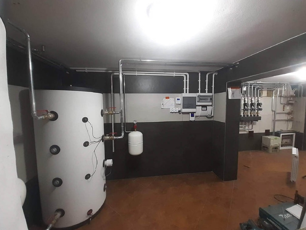Instalacja zbiornika buforowego ciepła z osprzętem w piwnicy w Rzozowie, widoczne liczne rury i zawory oraz panel sterowania.