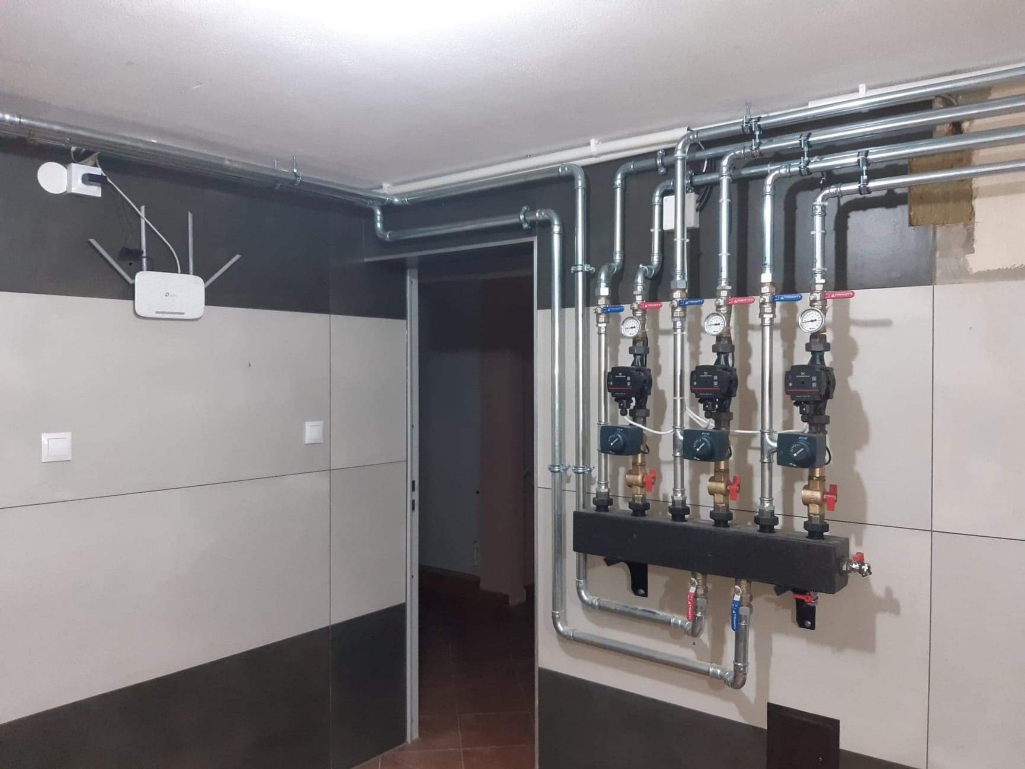 Rozdzielacz z trzema pompami obiegowymi zamontowany na ścianie w pomieszczeniu z instalacją rurową pod sufitem, widoczny router Wi-Fi.