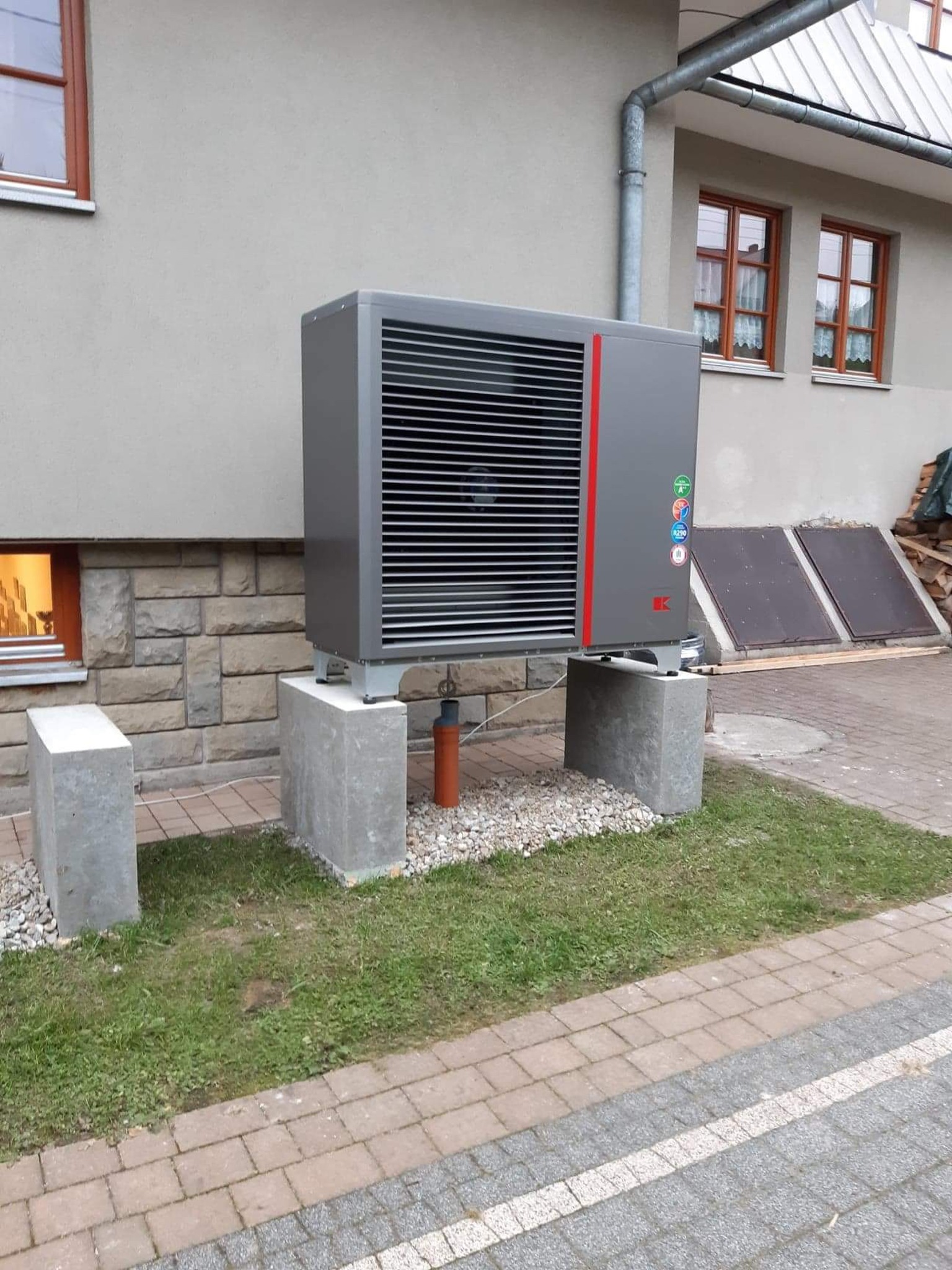 Szara pompa ciepła zamontowana na betonowych fundamentach obok domu z brązowymi oknami i widocznymi panelami solarnymi.