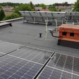 Widok z góry na płaski dach pokryty panelami słonecznymi, z ceglanym kominem i anteną telewizyjną w tle zabudowa miejska.