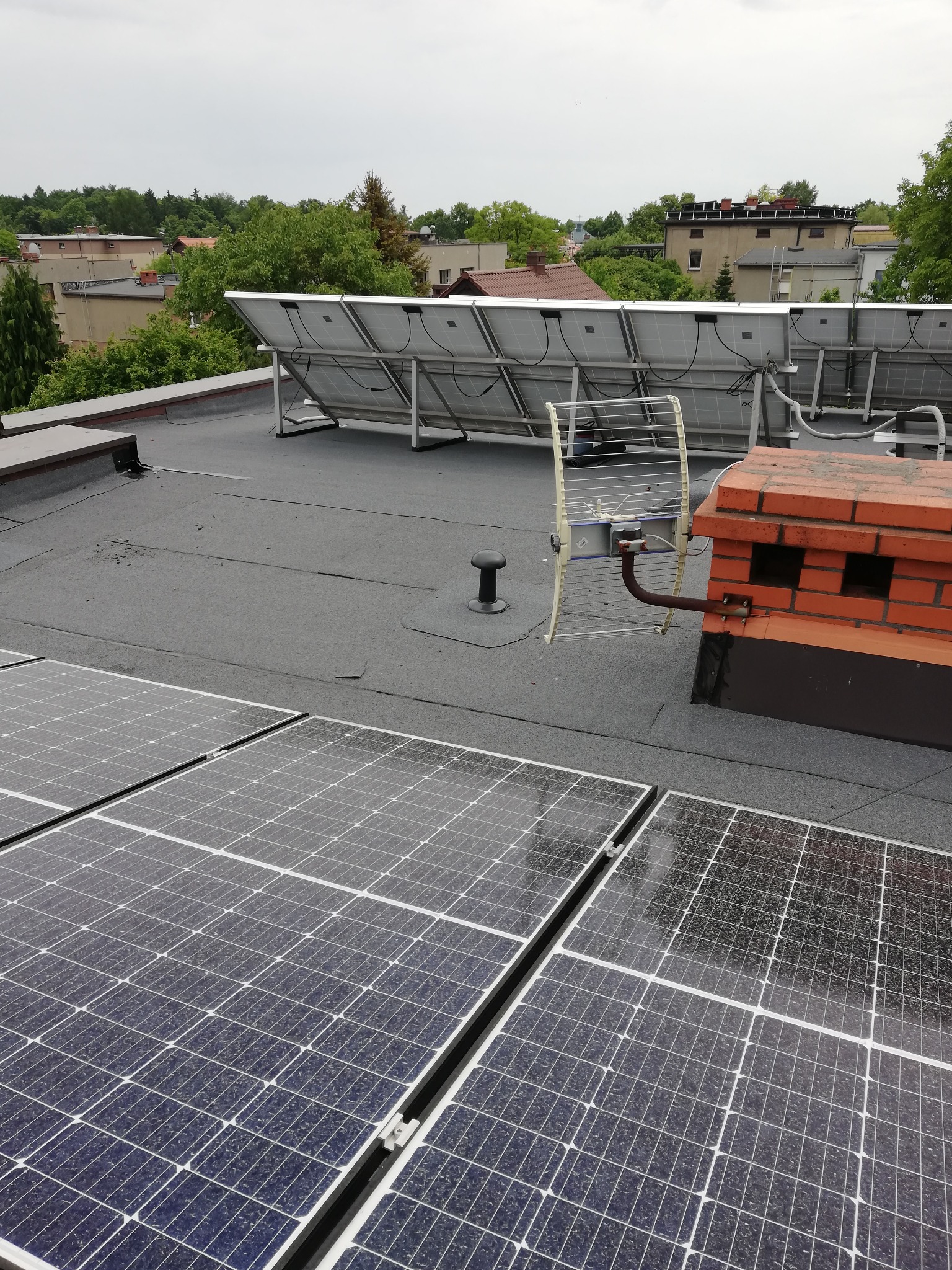Widok z góry na płaski dach pokryty panelami słonecznymi, z ceglanym kominem i anteną telewizyjną w tle zabudowa miejska.
