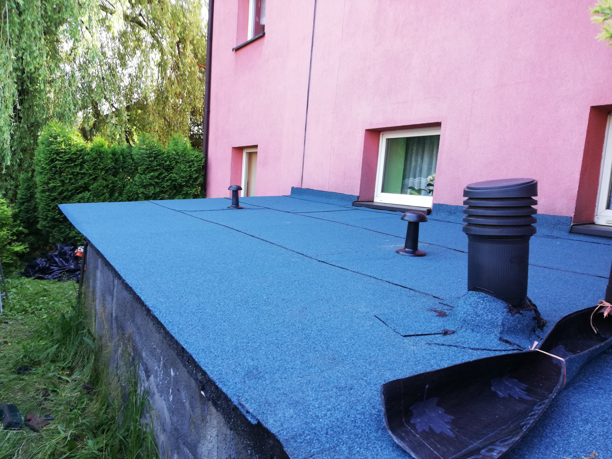 Płaski dach pokryty niebieską papą, z widocznymi kominkami wentylacyjnymi i fragmentem rynny z motywem liści, na tle różowej ściany budynku i zieleni.