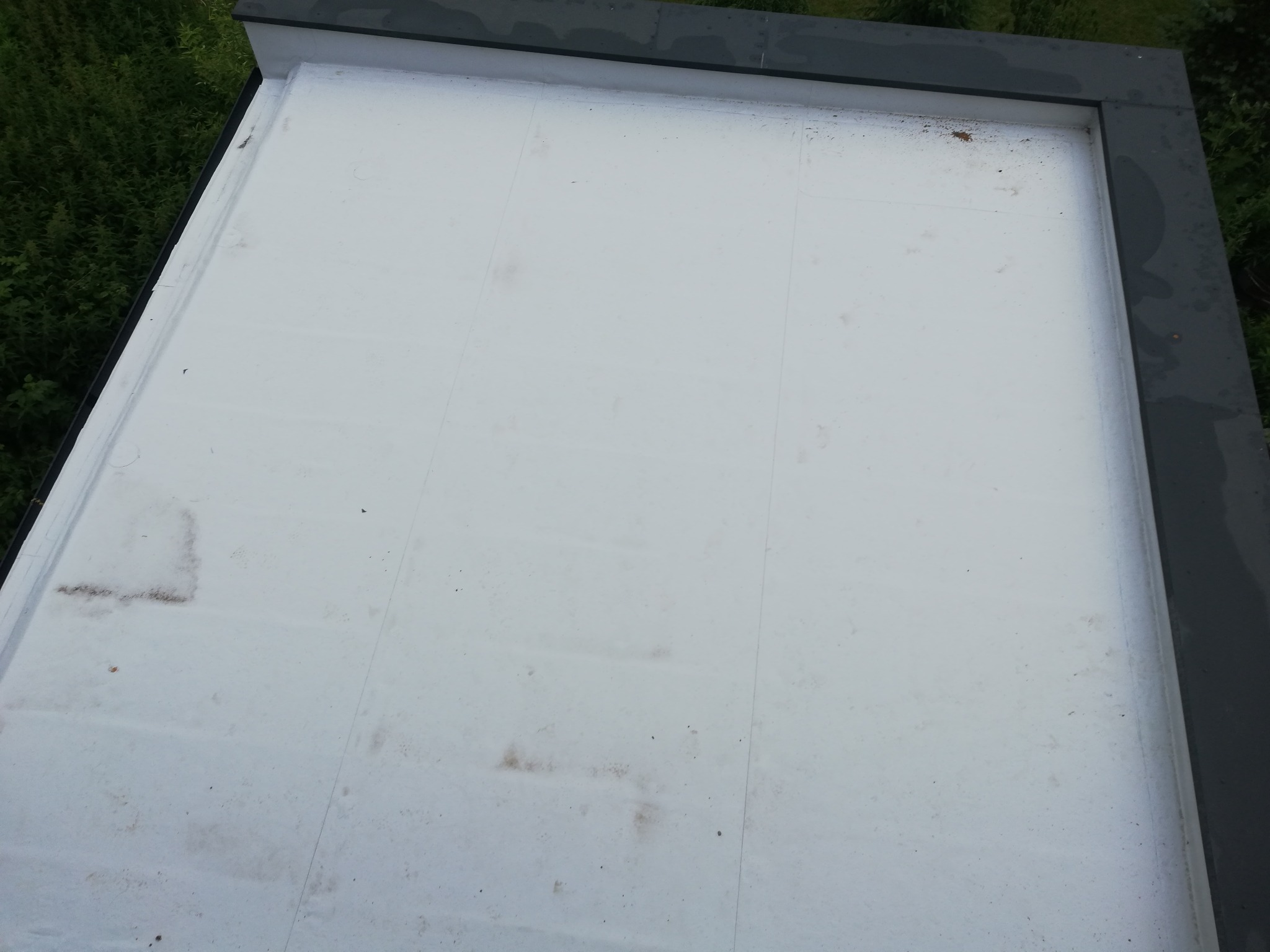 Widok z góry na płaski, biały dach pokryty papą termozgrzewalną, z widocznymi śladami zabrudzeń i zarysowaniami, obramowany szarym, metalowym profilem i otoczony zielenią drzew.