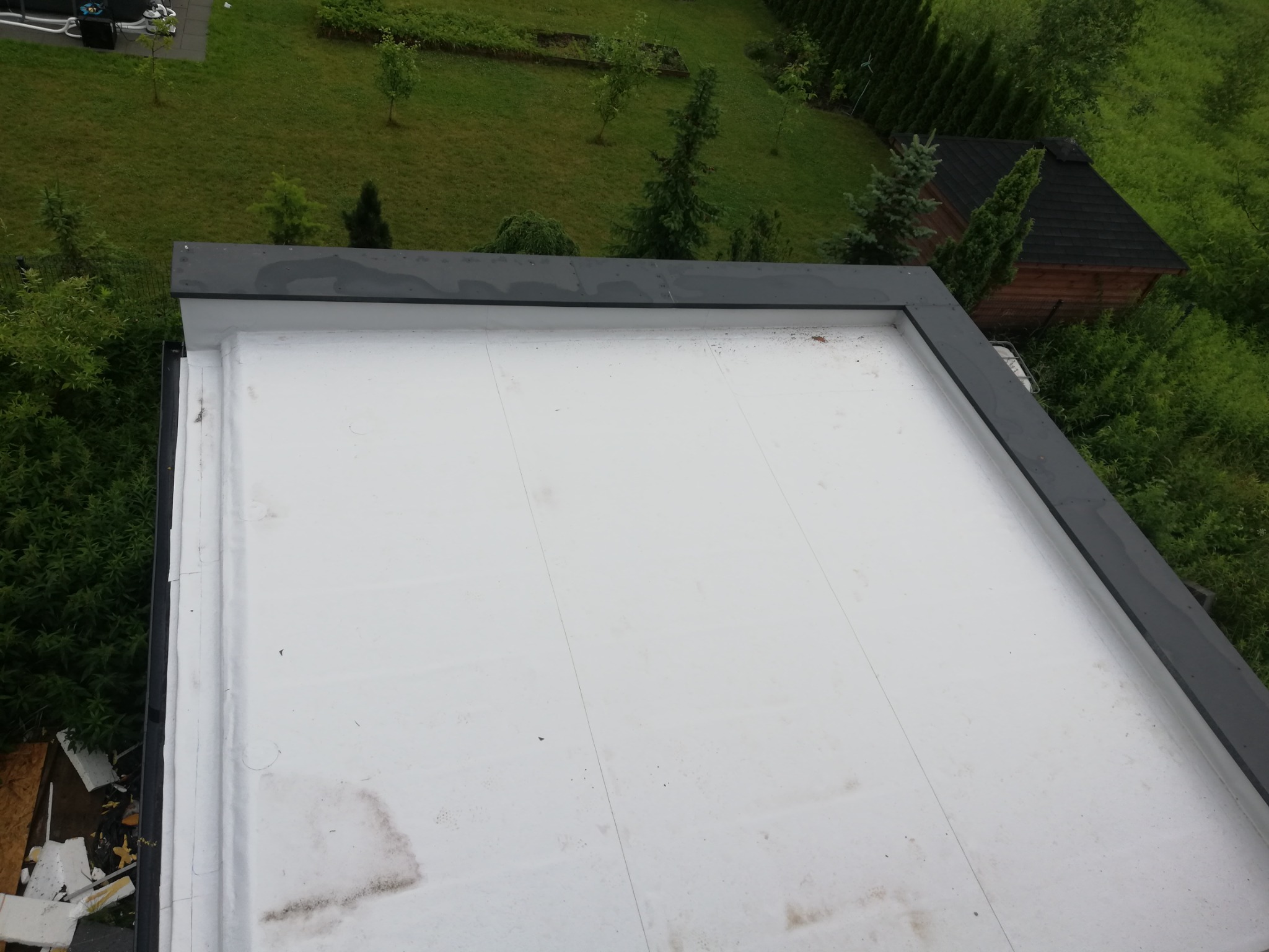 Biały, płaski dach z obróbką blacharską w kolorze grafitowym, widok z góry na tle zielonego ogrodu z domkiem narzędziowym.