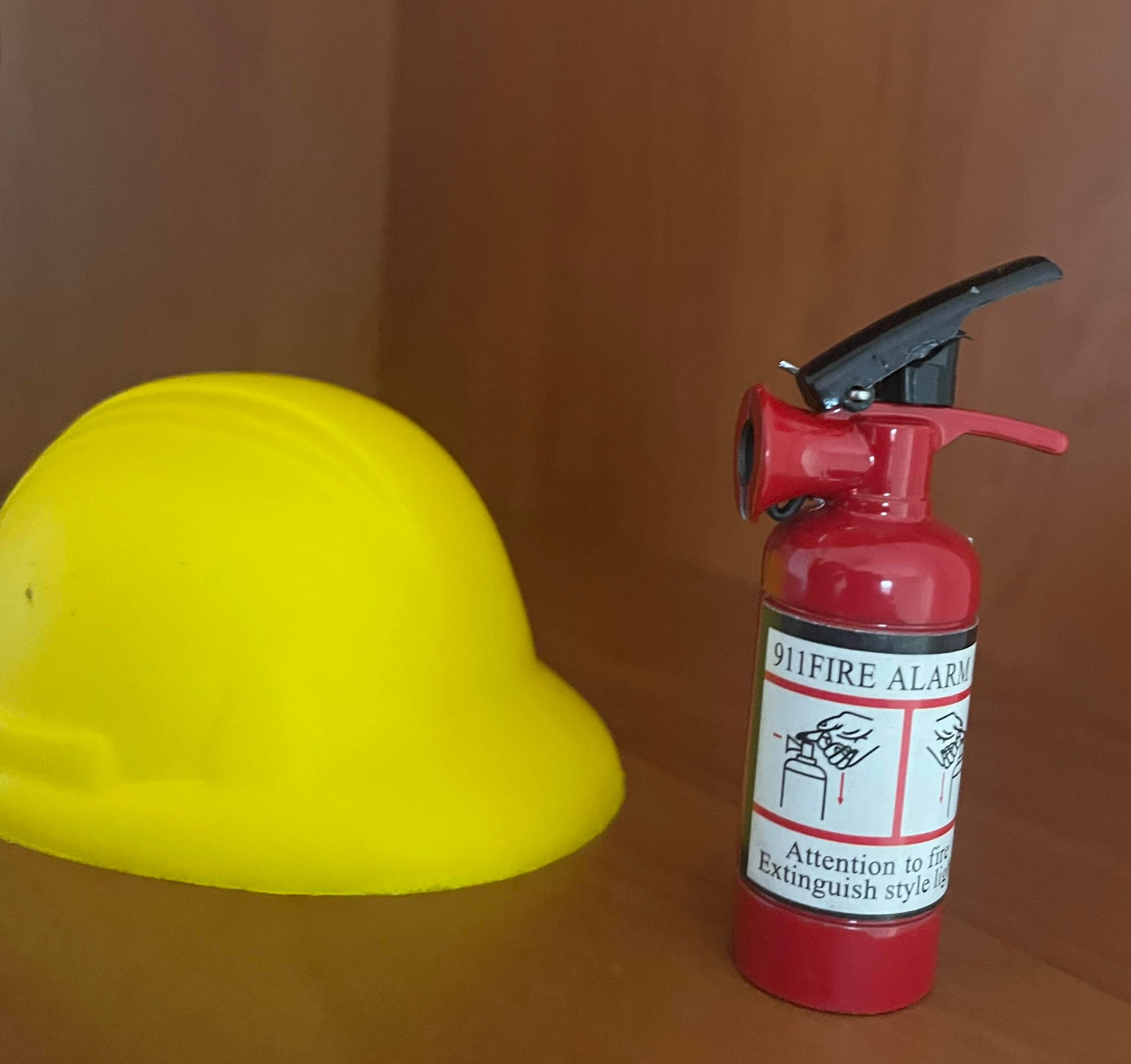 Żółty kask budowlany i miniaturowy czerwony gaśnica z napisem '911 Fire Alarm' na tle drewnianej ściany.