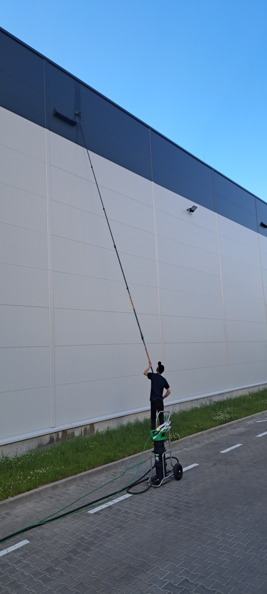 Pracownik na podeście, z długą lancą myjącą, czyści elewację hali przemysłowej z widocznymi smugami.