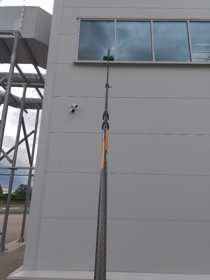 Mycie okien na wysokości budynku przemysłowego za pomocą teleskopowego kija z zieloną myjką, widoczna elewacja z szarego panelu i elementy konstrukcji stalowej.