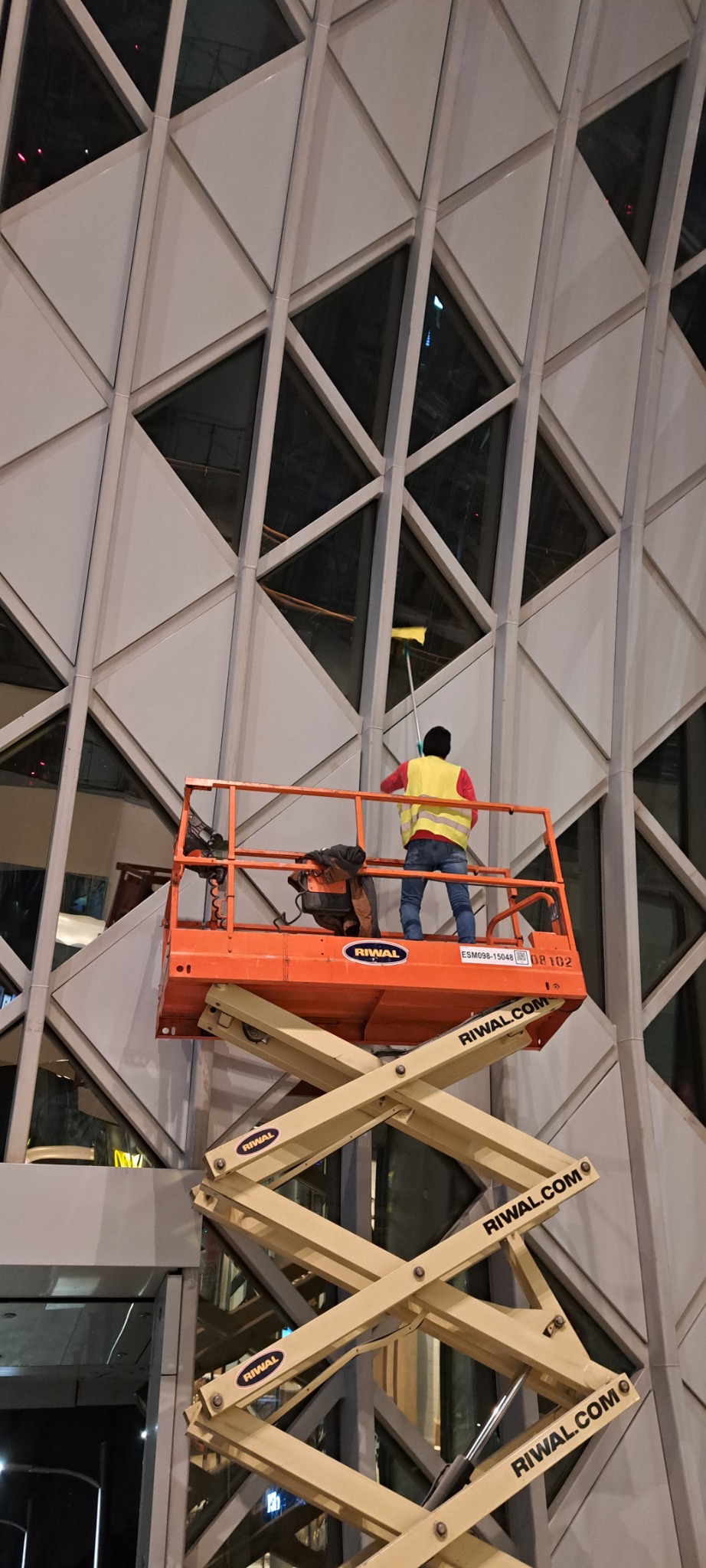 Pracownik w żółtej kamizelce na podnośniku Riwal czyści okna na wysokości w budynku o geometrycznej fasadzie.