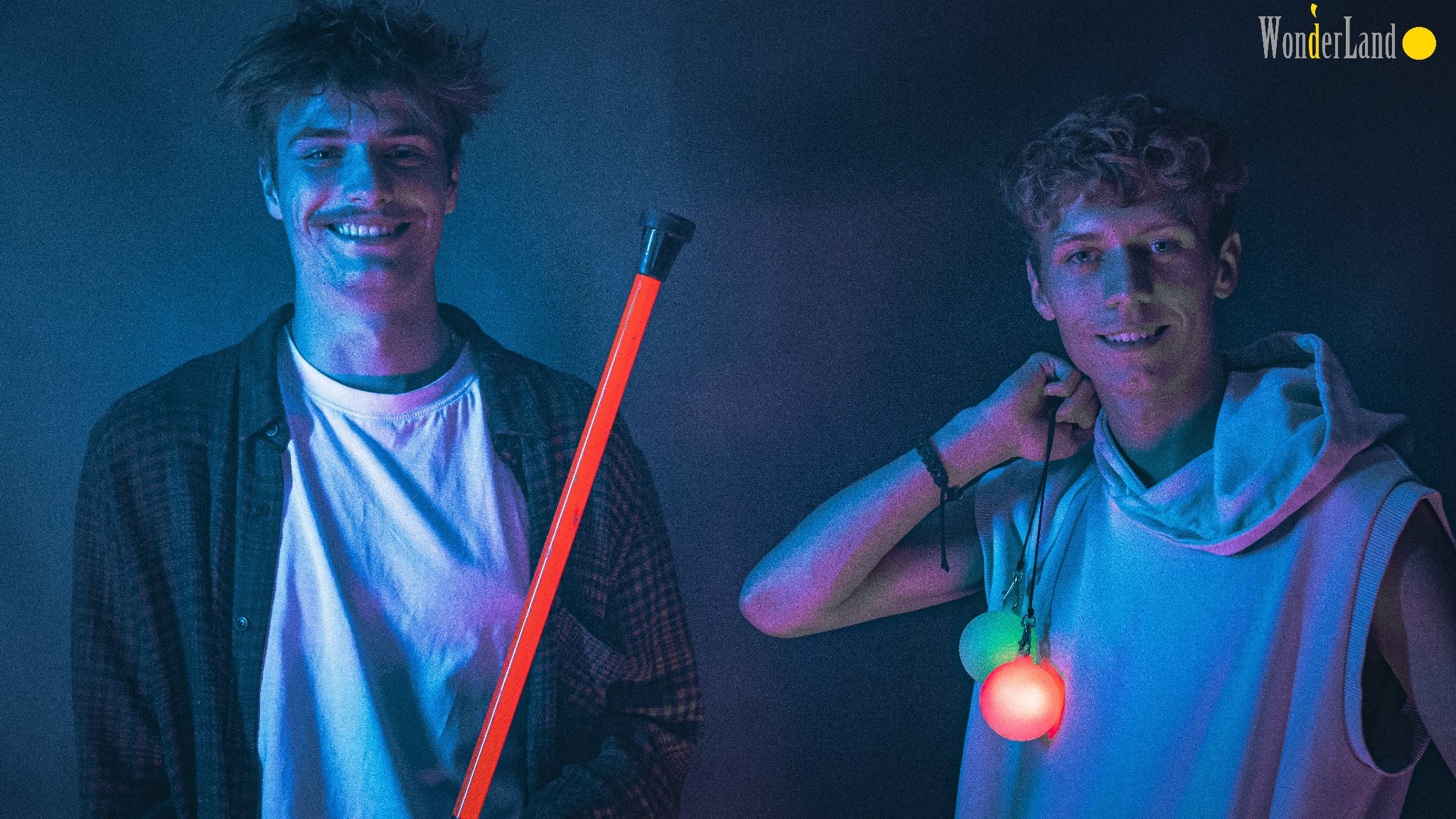 Dwóch uśmiechniętych młodych mężczyzn w niebieskim oświetleniu, jeden trzyma świecącą, pomarańczową laskę, drugi ma świecące kule na szyi, w tle logo WonderLand.