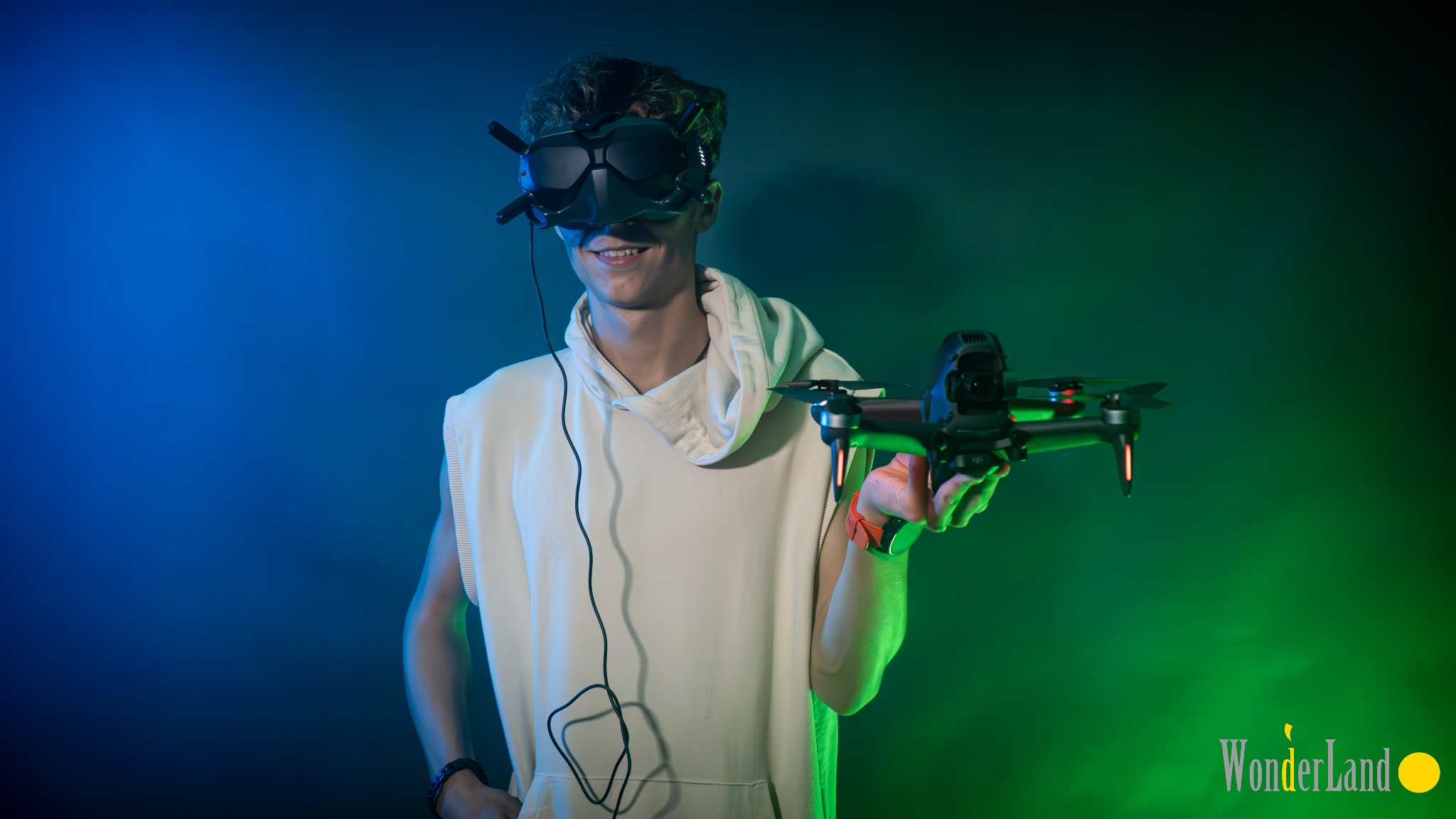 Uśmiechnięty nastolatek w goglach VR trzymający drona na tle niebiesko-zielonego oświetlenia studyjnego.