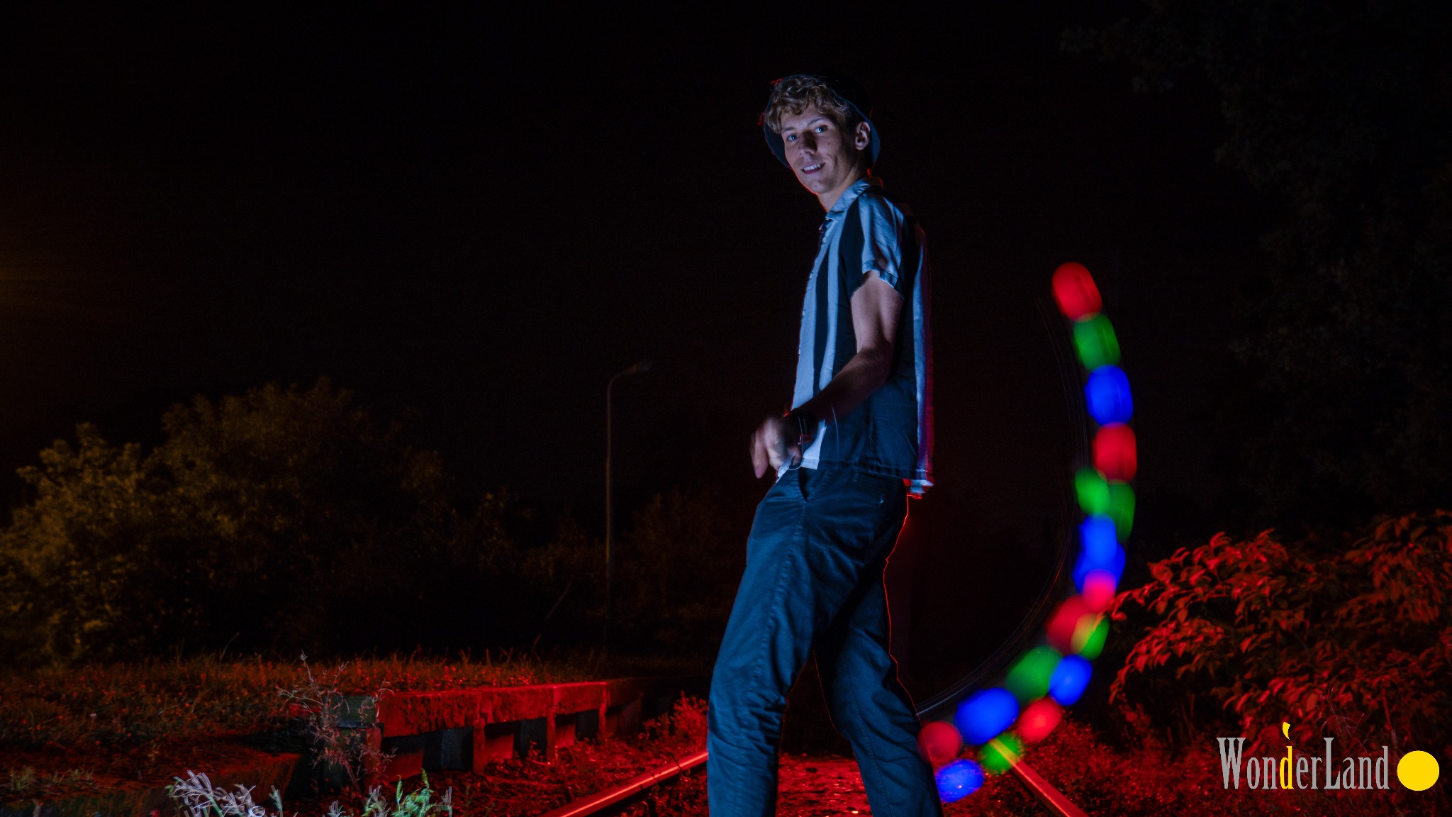 Młody mężczyzna w czarno-białej koszulce, stojący na torach kolejowych nocą, kręci kolorowymi światłami LED, tworząc efektowny, rozmyty okrąg.