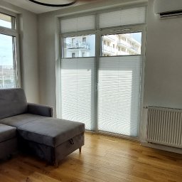 BEAUTY DESIGN J. OŻÓG - Jasne wnętrze salonu z szarym narożnikiem i oknami przesłoniętymi białymi plisami, widok na budynki za oknem.