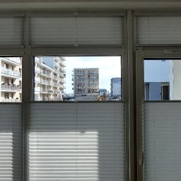 BEAUTY DESIGN J. OŻÓG - Zamknięte, białe plisy okienne w trzyskrzydłowym oknie z widokiem na nowoczesne budynki mieszkalne.