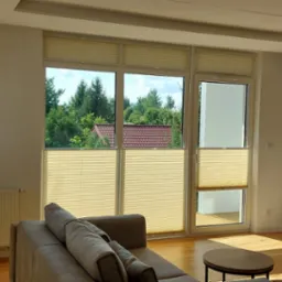 Jasne plisy okienne w nowoczesnym salonie z widokiem na zieleń, częściowo zasłonięte okno balkonowe.