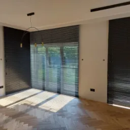 Nowoczesne wnętrze z drewnianą podłogą w jodełkę i trzema oknami zasłoniętymi ciemnymi żaluzjami poziomymi, przez które widać zieleń na zewnątrz, oświetlone światłem słonecznym.