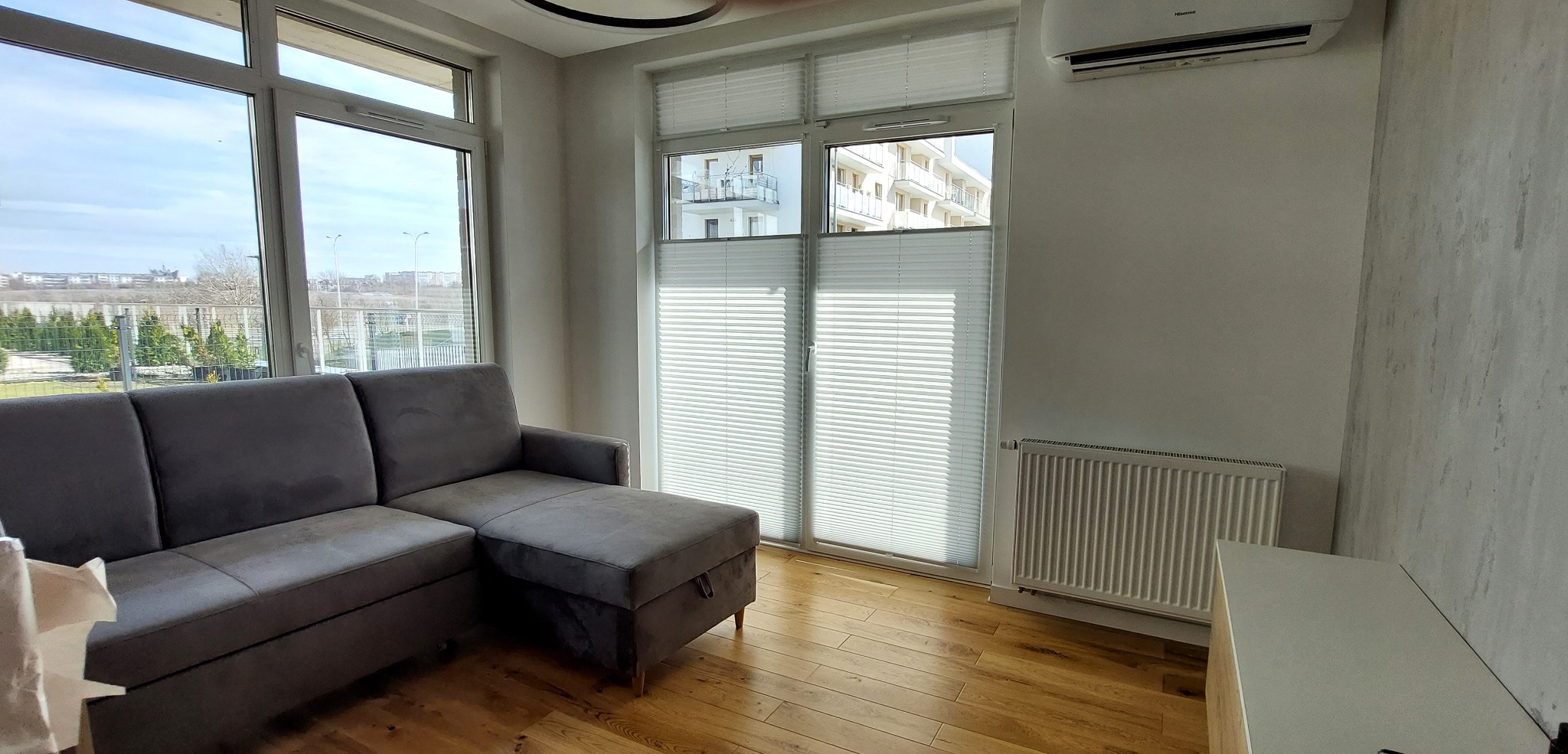 Jasne wnętrze salonu z szarym narożnikiem i oknami przesłoniętymi białymi plisami, widok na budynki za oknem.