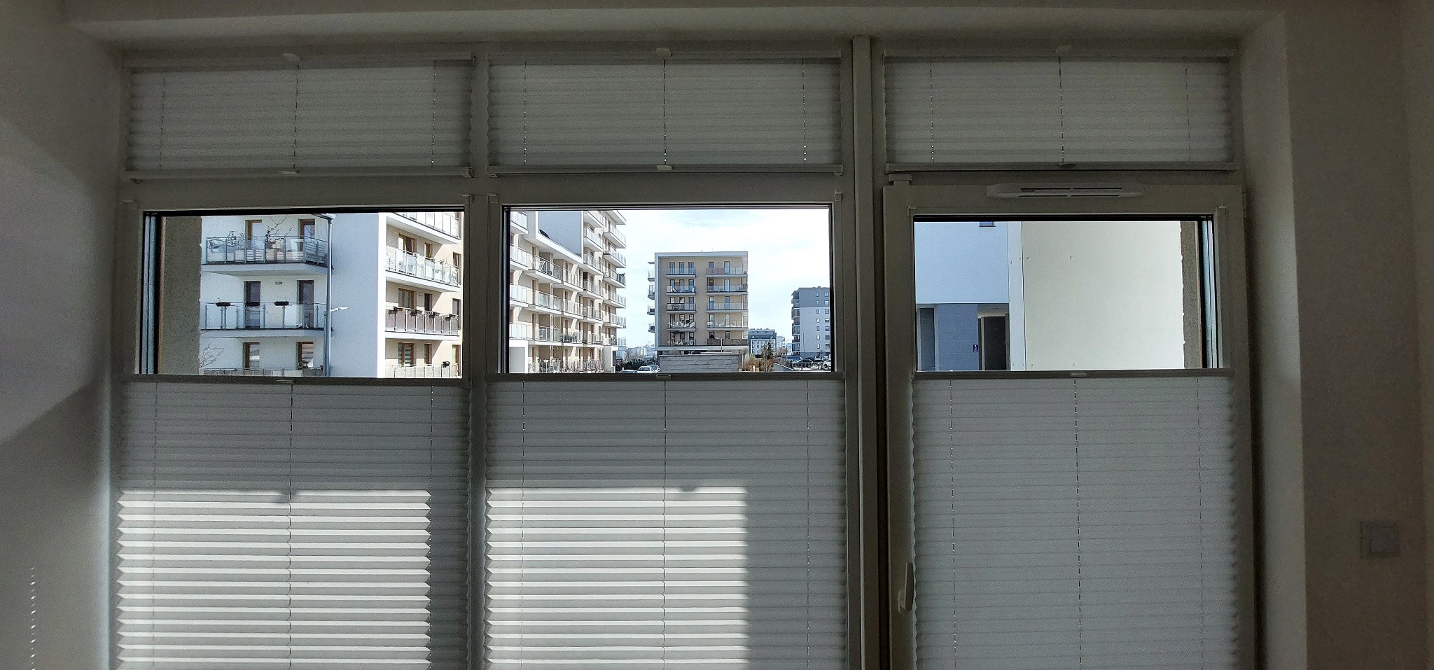 Zamknięte, białe plisy okienne w trzyskrzydłowym oknie z widokiem na nowoczesne budynki mieszkalne.