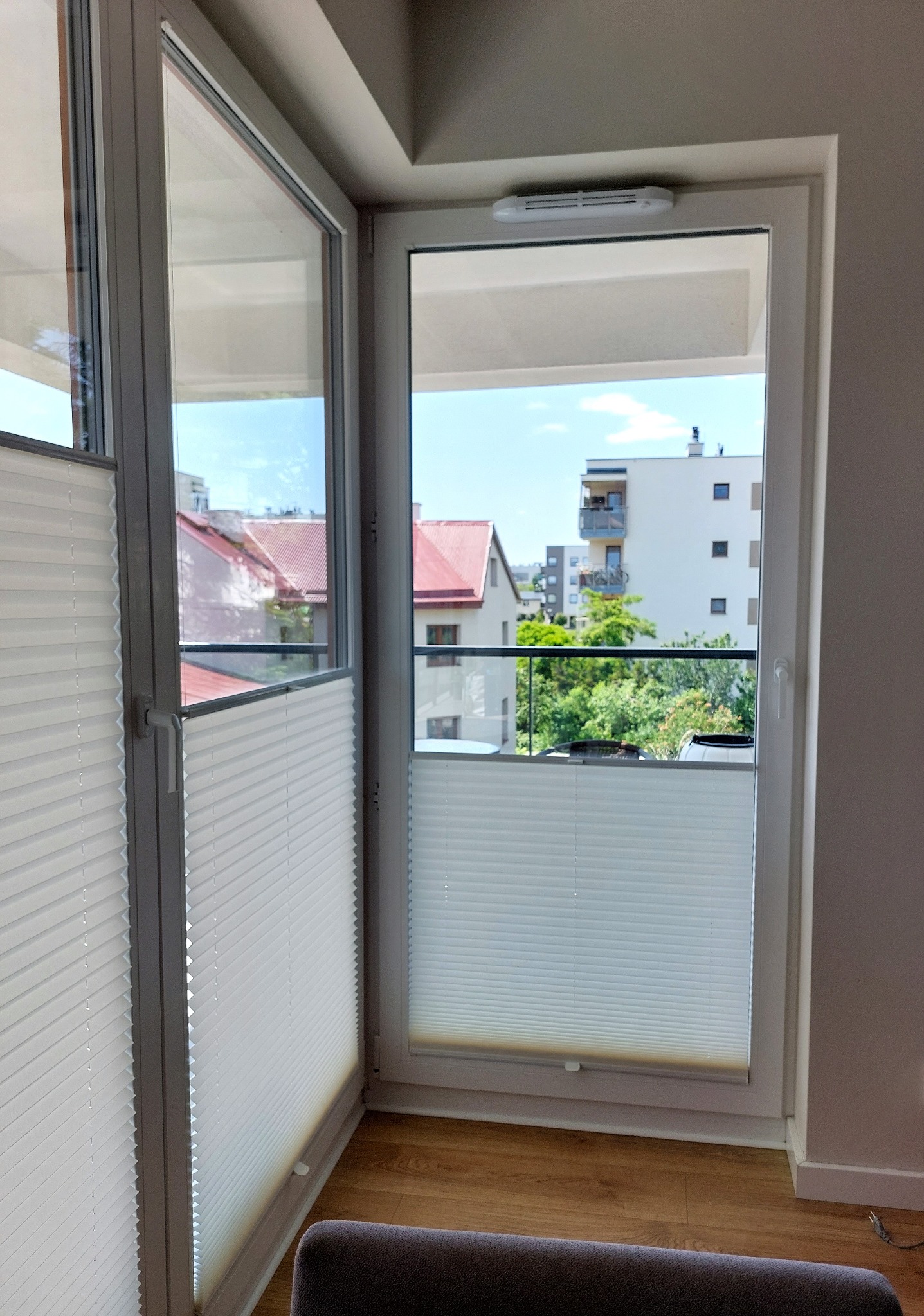Białe plisy okienne zamontowane w oknie balkonowym, widok na budynki mieszkalne i zieleń na zewnątrz.