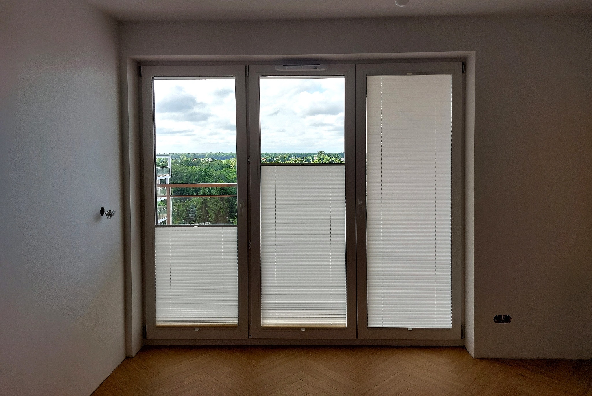 Trzyszybowe okno z zamontowanymi białymi plisami, częściowo zasłoniętymi, widok na zieleń i budynki z balkonu, parkiet na podłodze, surowe ściany w trakcie wykańczania.