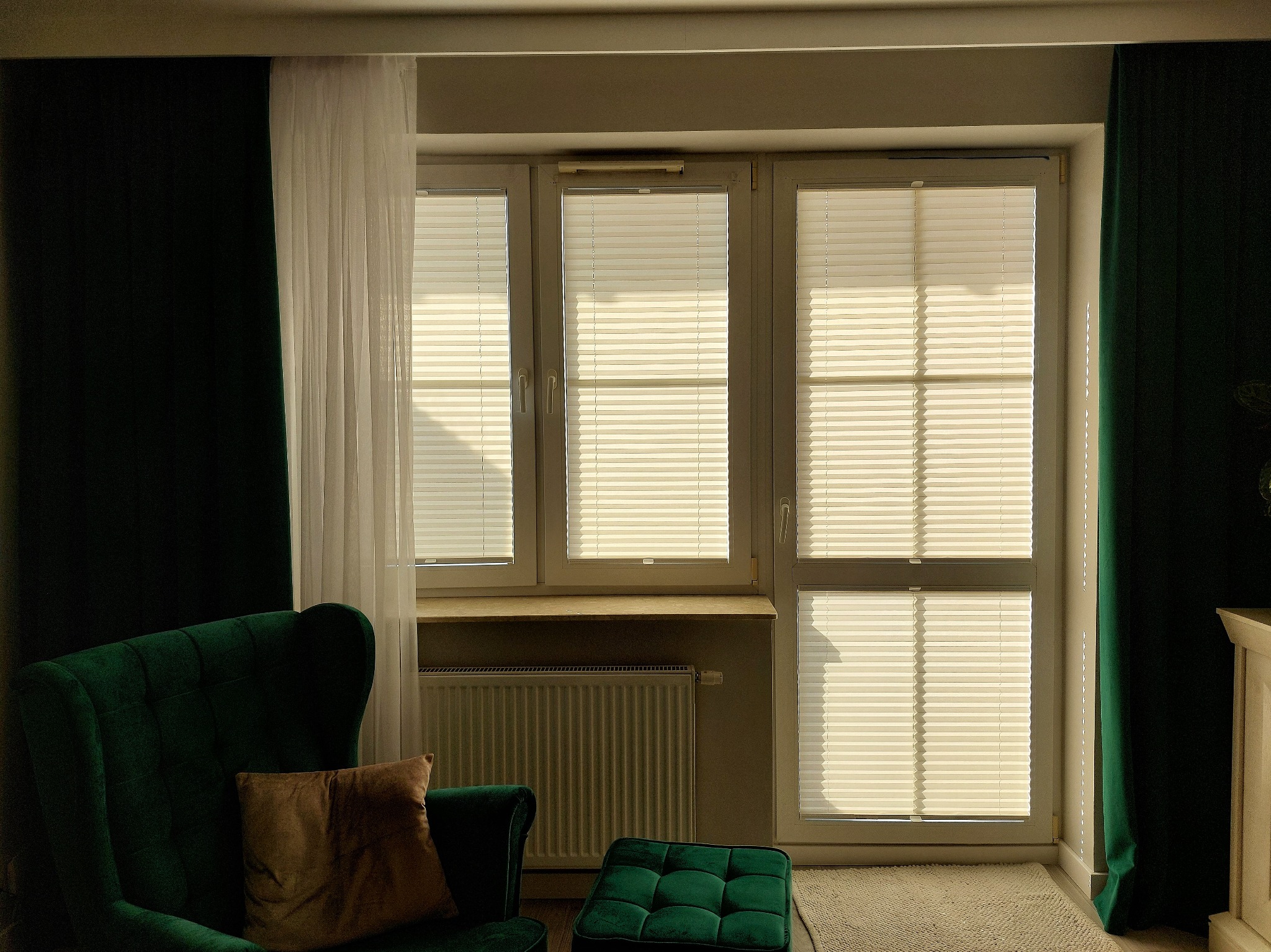 Trzy okna z zamontowanymi plisami w jasnym odcieniu, widoczne z wnętrza pokoju z zielonym fotelem i pufą, zasłonięte zielonymi i białymi zasłonami.