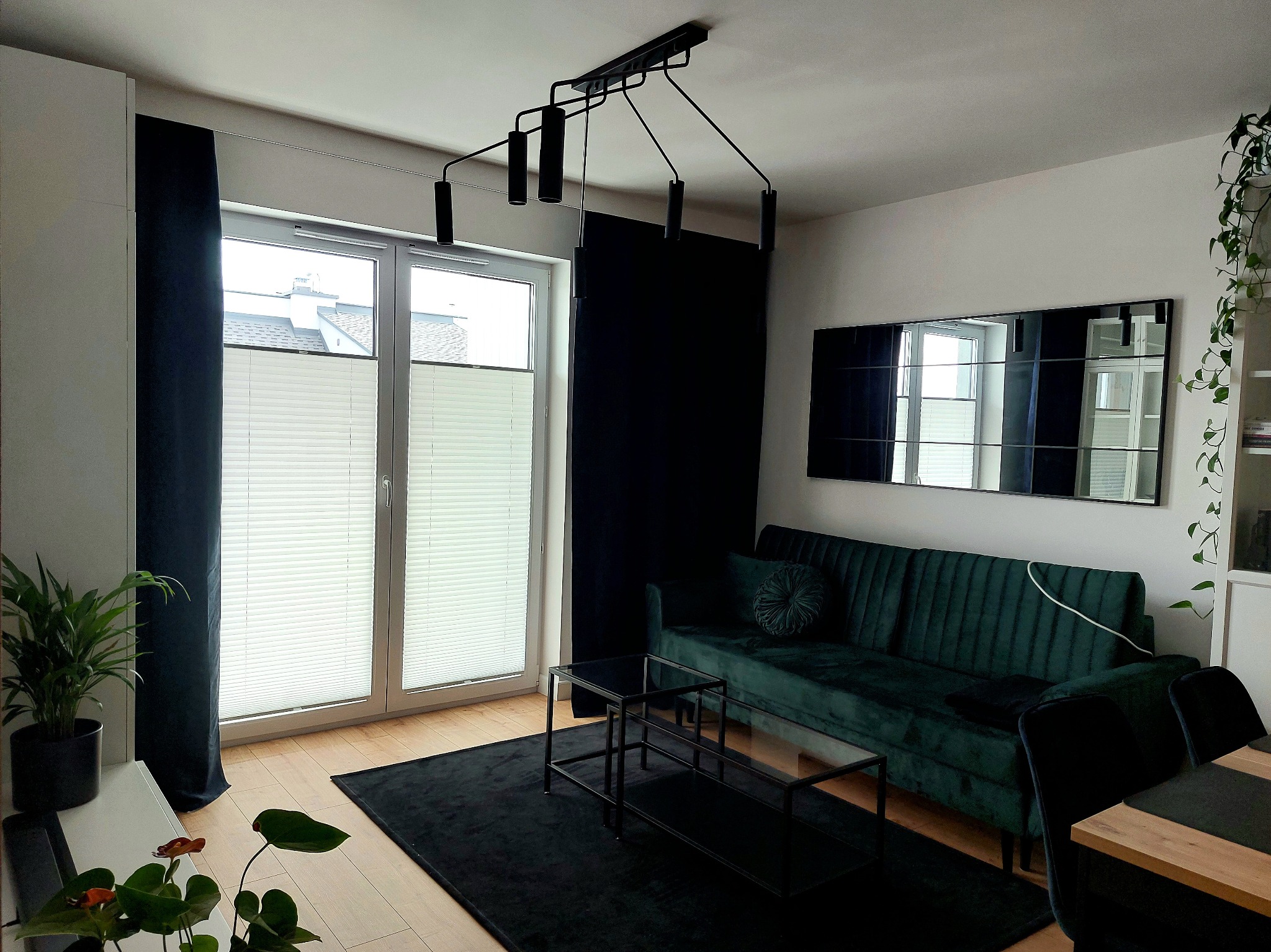 Salon z zieloną sofą, czarnym dywanem i plisowanymi roletami na oknach, oświetlony nowoczesną lampą sufitową.