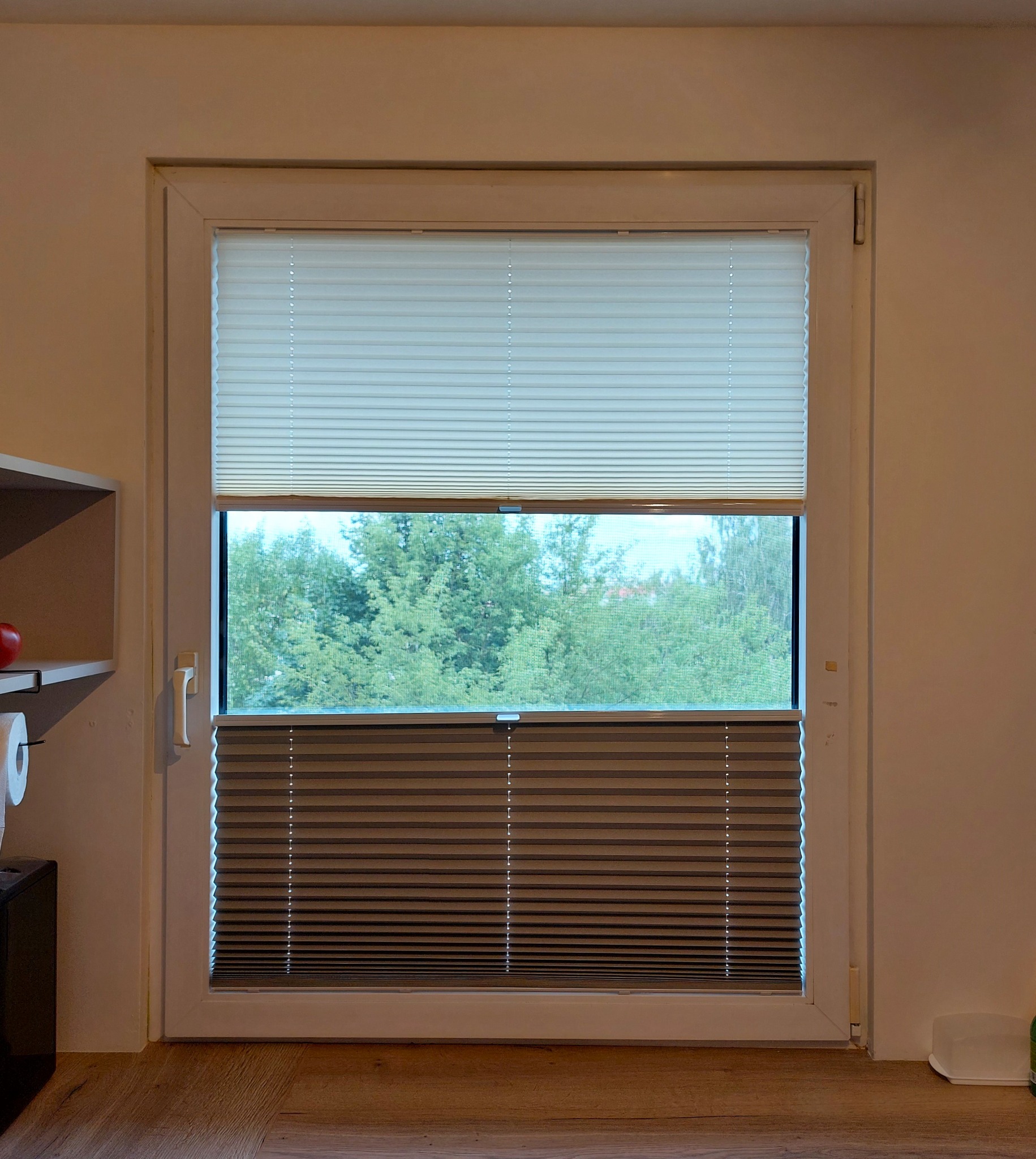 Okno z zamontowanymi plisami: górna część biała, dolna brązowa, widok na zieleń za oknem.