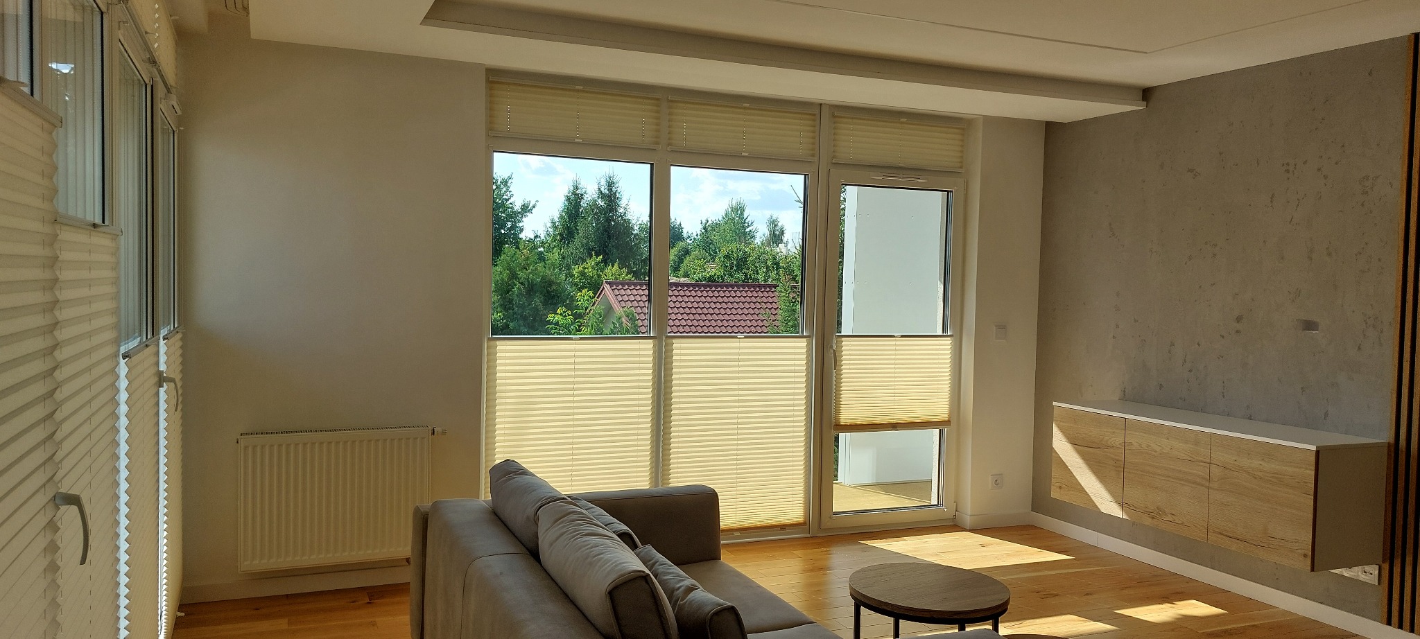 Jasne plisy okienne w nowoczesnym salonie z widokiem na zieleń, częściowo zasłonięte okno balkonowe.