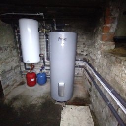 OFF GRID SPÓŁKA Z OGRANICZONĄ ODPOWIEDZIALNOŚCIĄ - Dwa zbiorniki buforowe w piwnicy z ceglanymi ścianami, jeden biały, drugi szary z logo Ferroli, obok czerwony i niebieski zbiornik wyrównawczy.