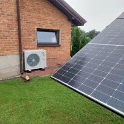 Zewnętrzna jednostka pompy ciepła Rotenso Aquami obok ceglanego budynku, w tle panele fotowoltaiczne z kroplami deszczu.