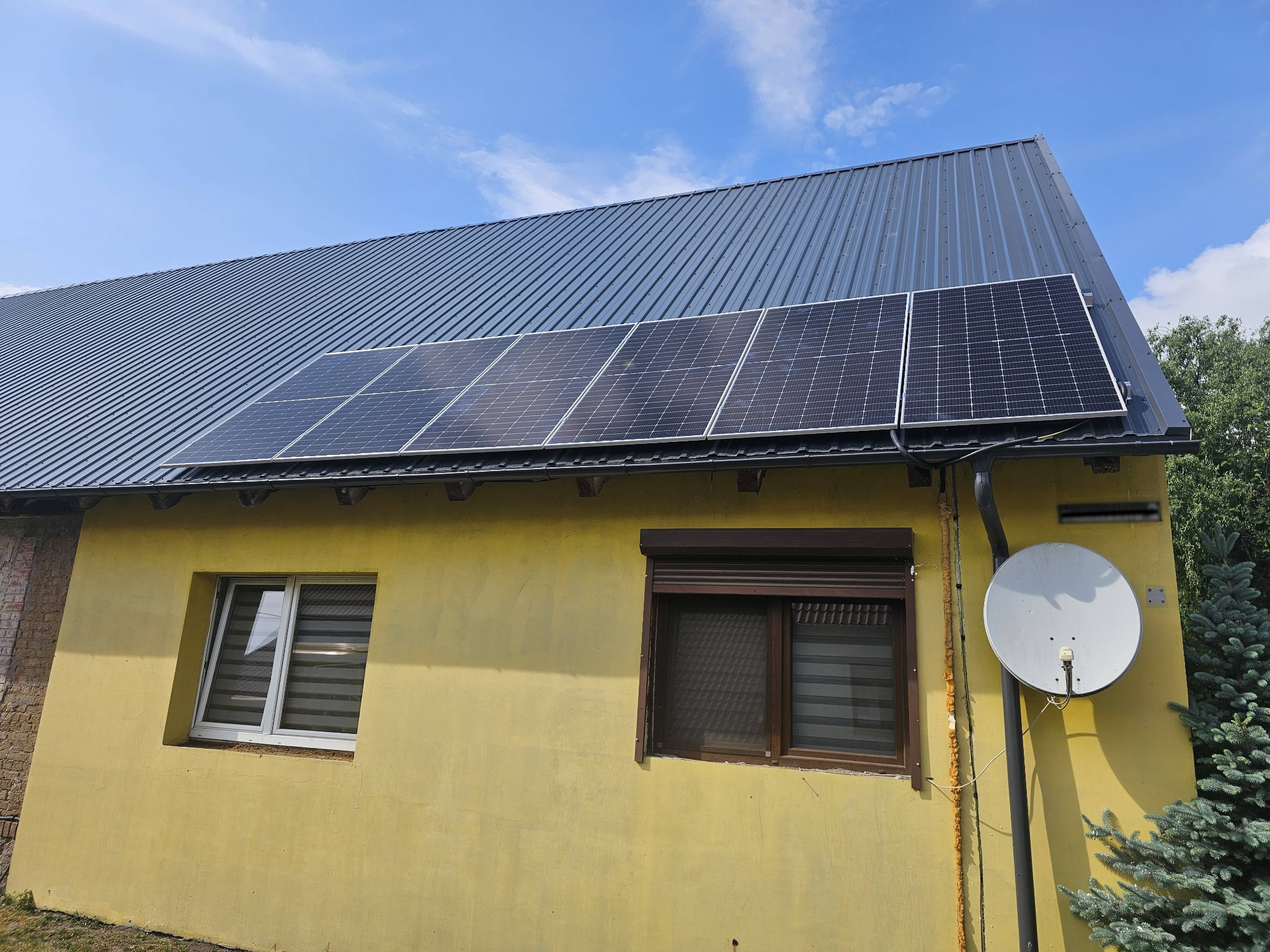 Instalacja paneli słonecznych na dachu domu z blachy trapezowej, żółta elewacja, widoczna antena satelitarna i okna z roletami zewnętrznymi.