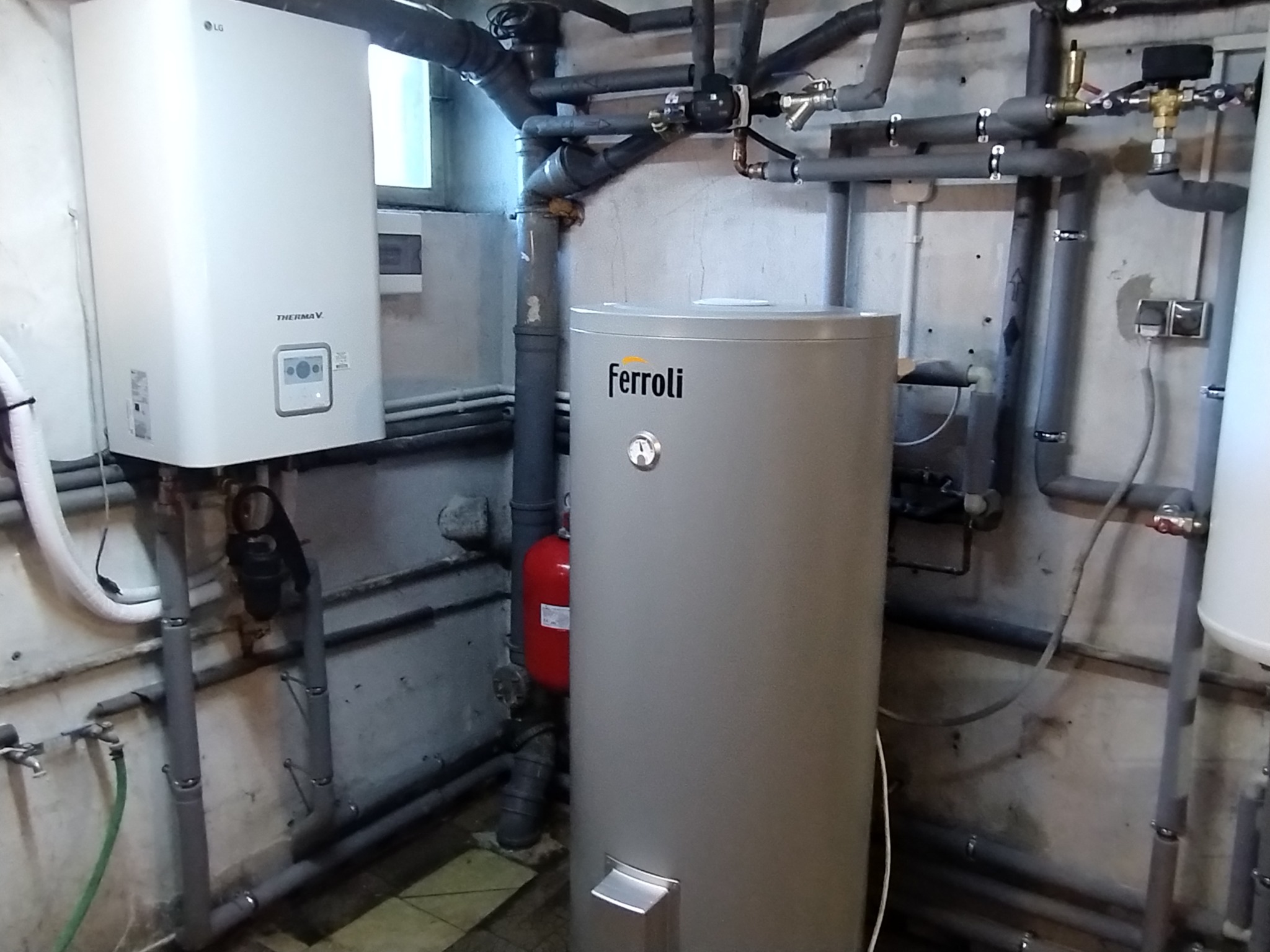 Kotłownia z dwoma zbiornikami: biały LG Therma V wiszący na ścianie i szary Ferroli stojący na podłodze, otoczona siecią rur i zaworów.