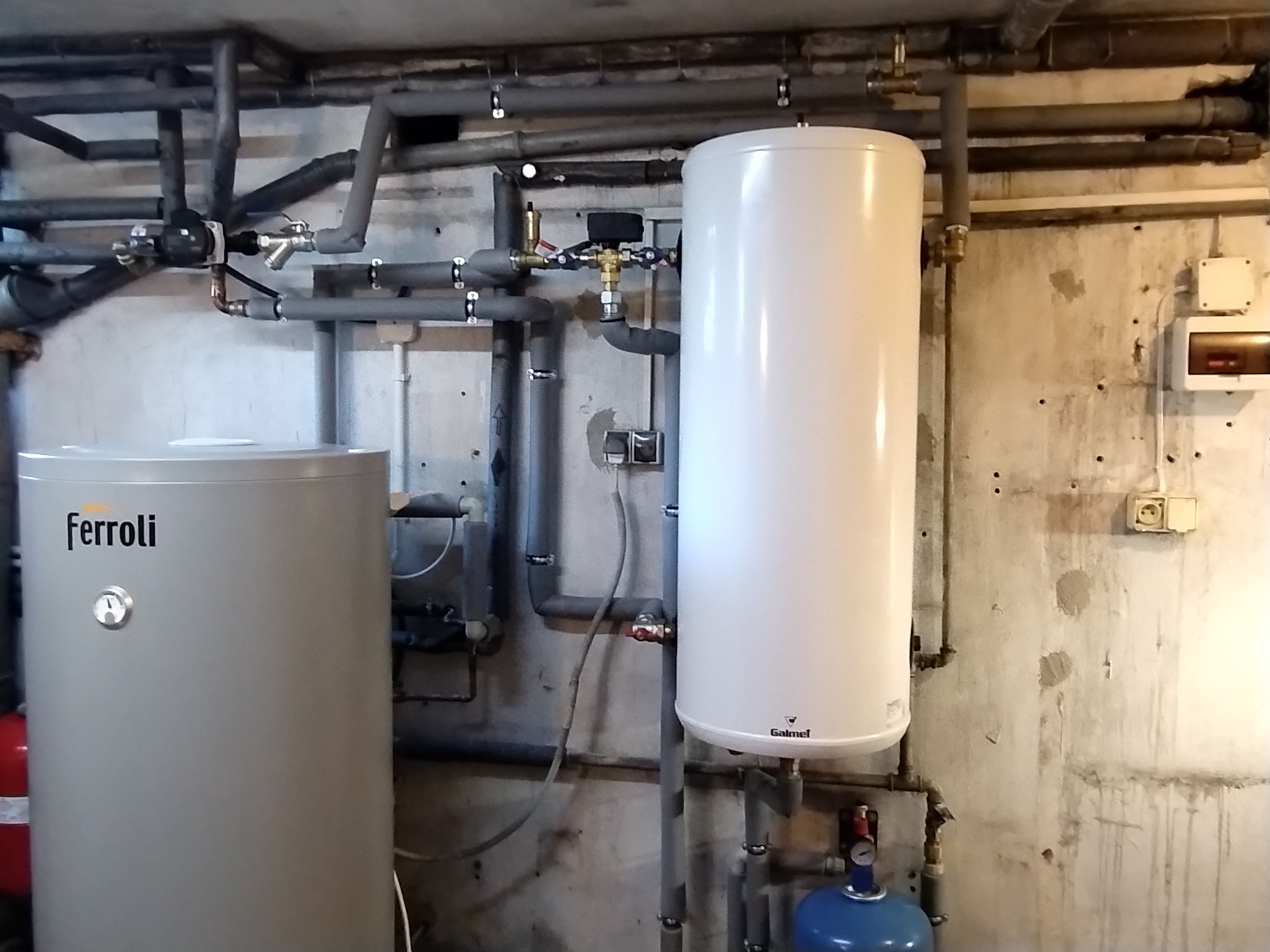 Dwa zbiorniki na wodę, jeden szary marki Ferroli z widocznym termometrem, drugi biały marki Galmet, połączone rurami w piwnicy z surowymi ścianami.