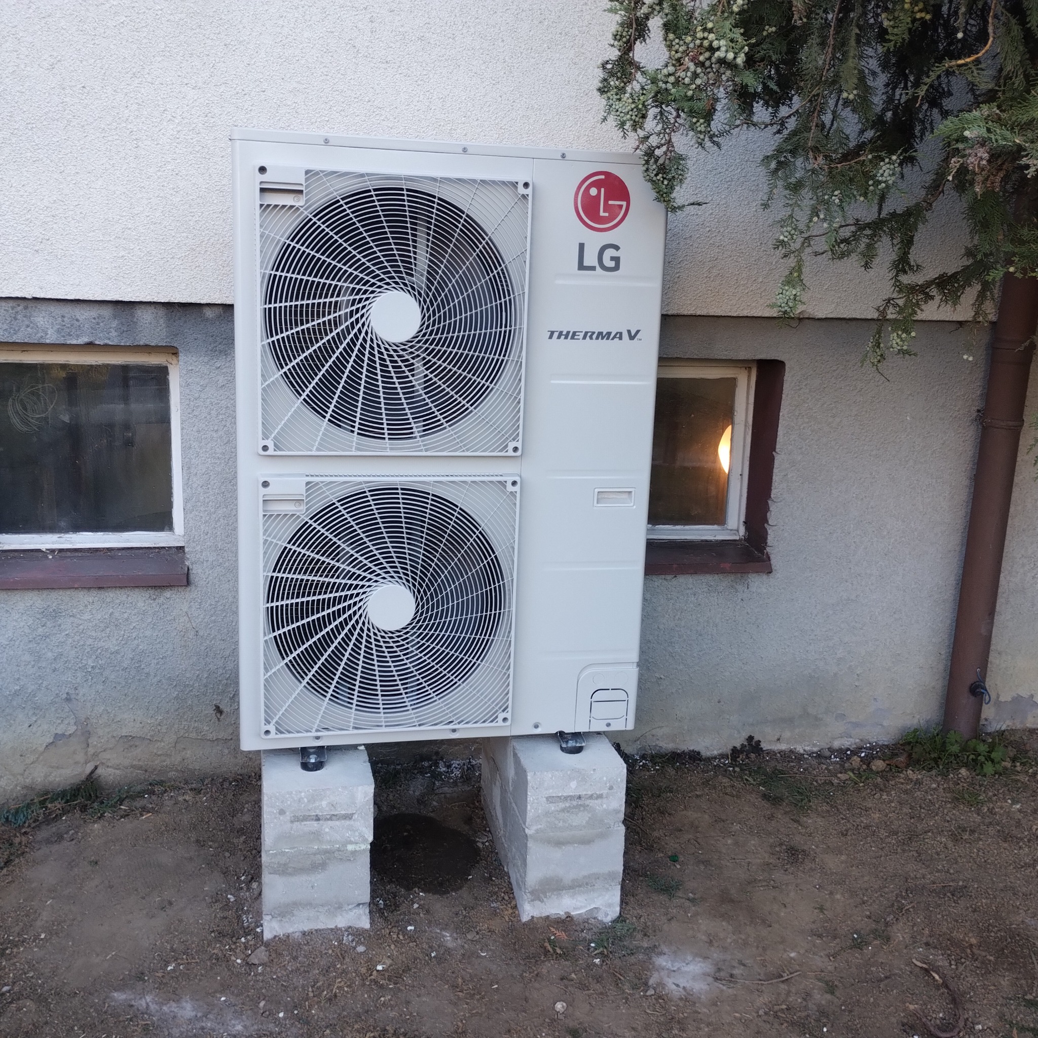 Biała jednostka zewnętrzna pompy ciepła LG Therma V, umieszczona na betonowych bloczkach przed budynkiem z oknami i rynną.