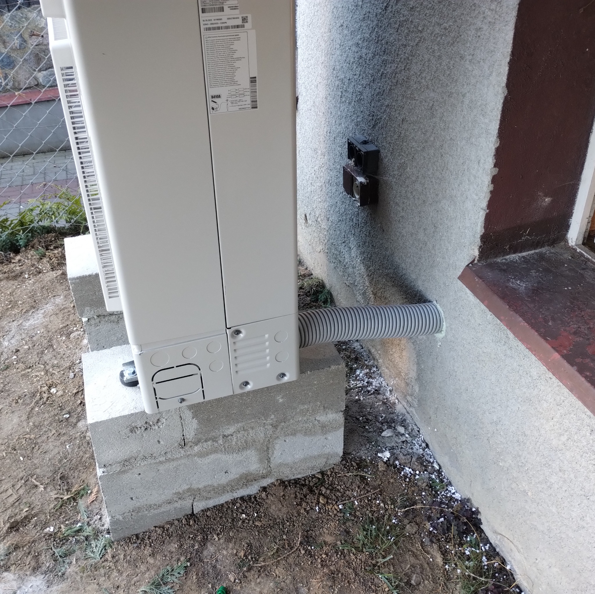 Zewnętrzna jednostka klimatyzacji zamontowana na betonowych bloczkach, z widocznym przewodem odprowadzającym skropliny przez ścianę budynku.