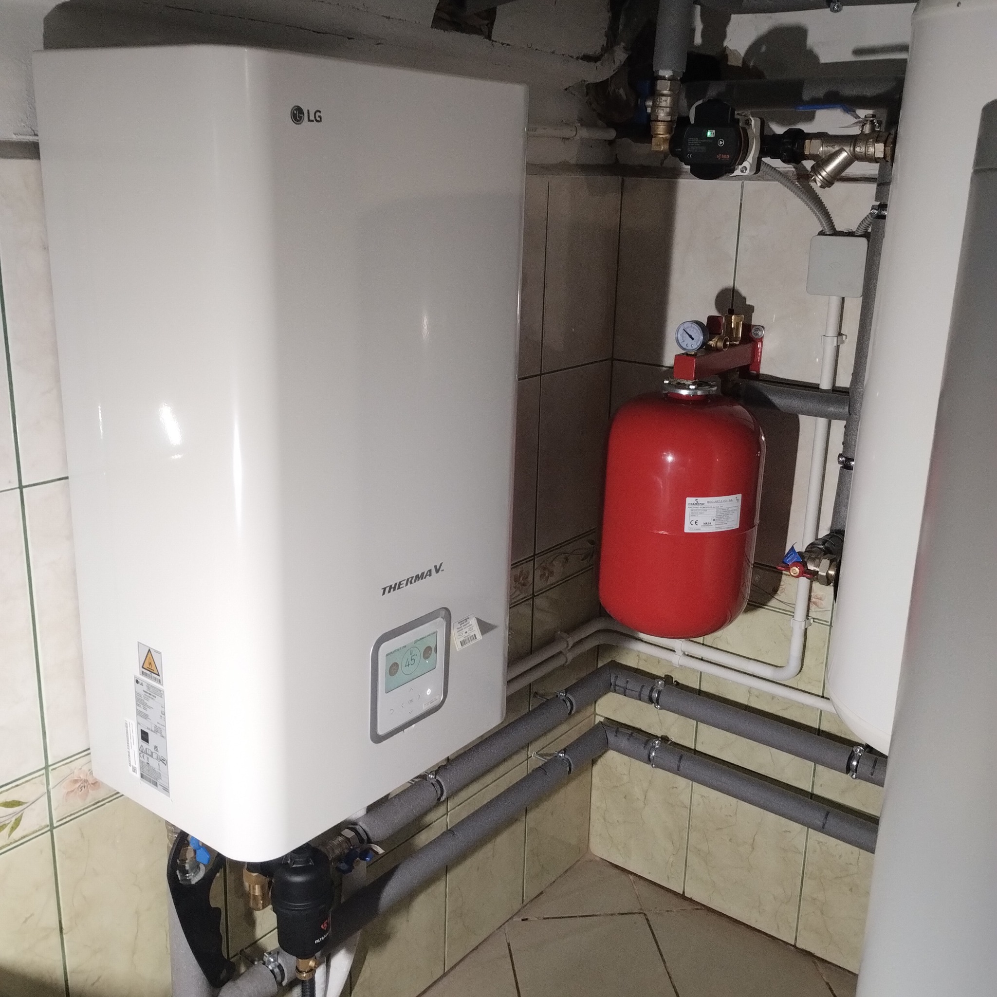 Biała pompa ciepła LG Therma V zamontowana w kotłowni, widoczne rury w szarej izolacji, czerwony zbiornik wyrównawczy i fragment białego zasobnika ciepłej wody.