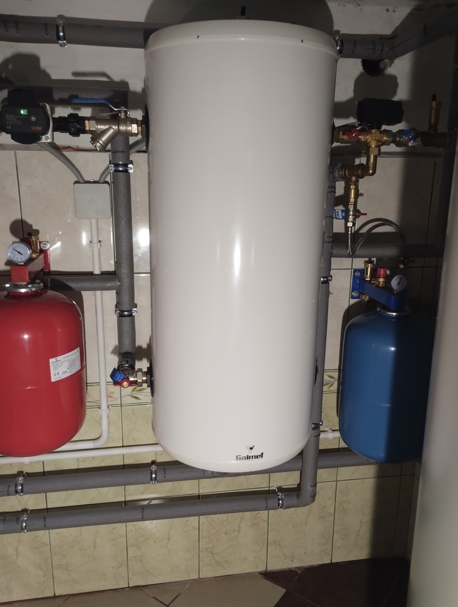 Instalacja z białym zasobnikiem ciepłej wody marki Galmet, czerwonym i niebieskim zbiornikiem wyrównawczym, rurami w szarej izolacji oraz zaworami i manometrami na tle beżowych płytek.