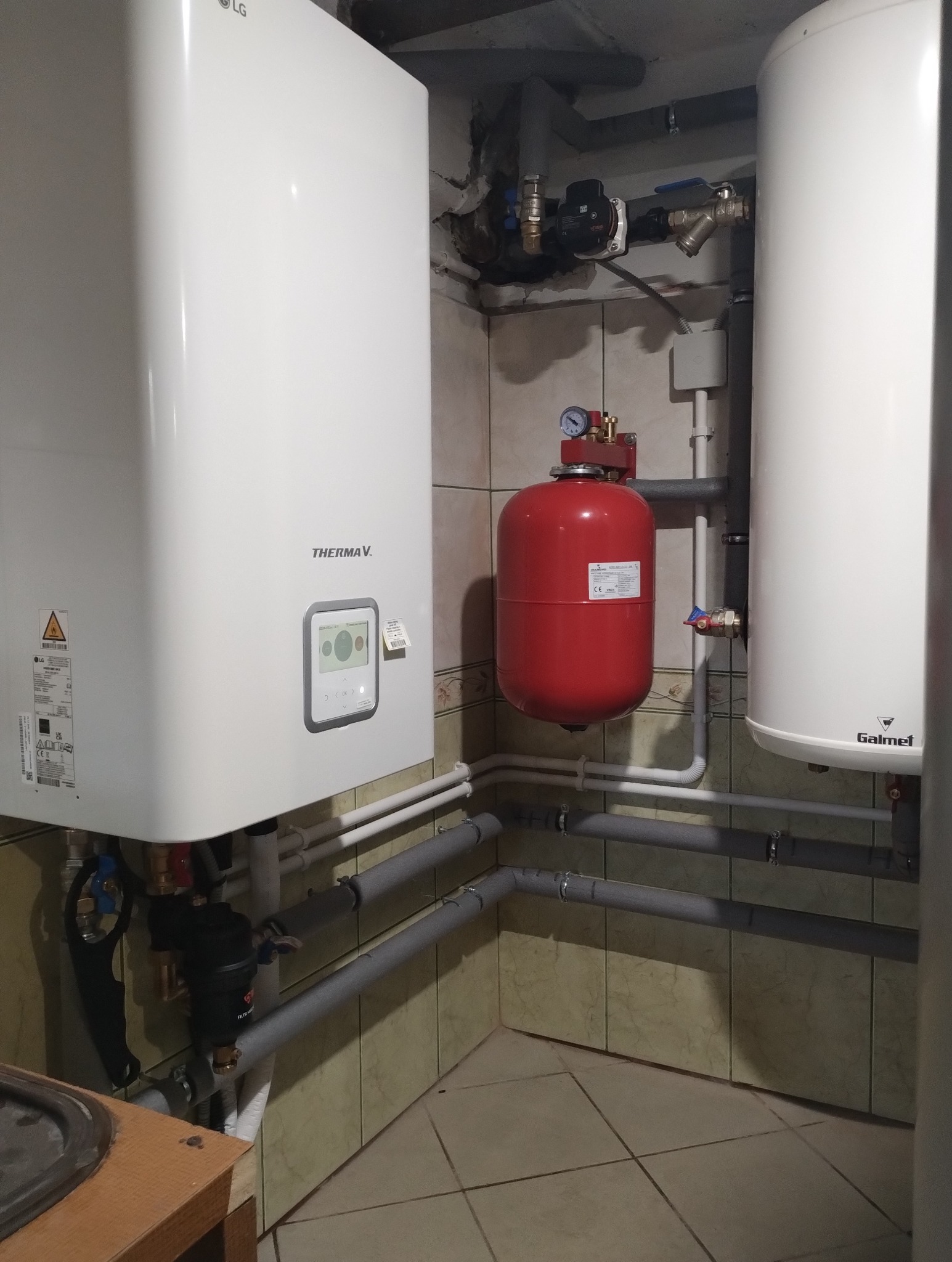 Instalacja kotła grzewczego THERMA V. z czerwonym zbiornikiem wyrównawczym i białym zasobnikiem ciepłej wody Galmet, widoczne rury instalacyjne i armatura, w tle kafelki.