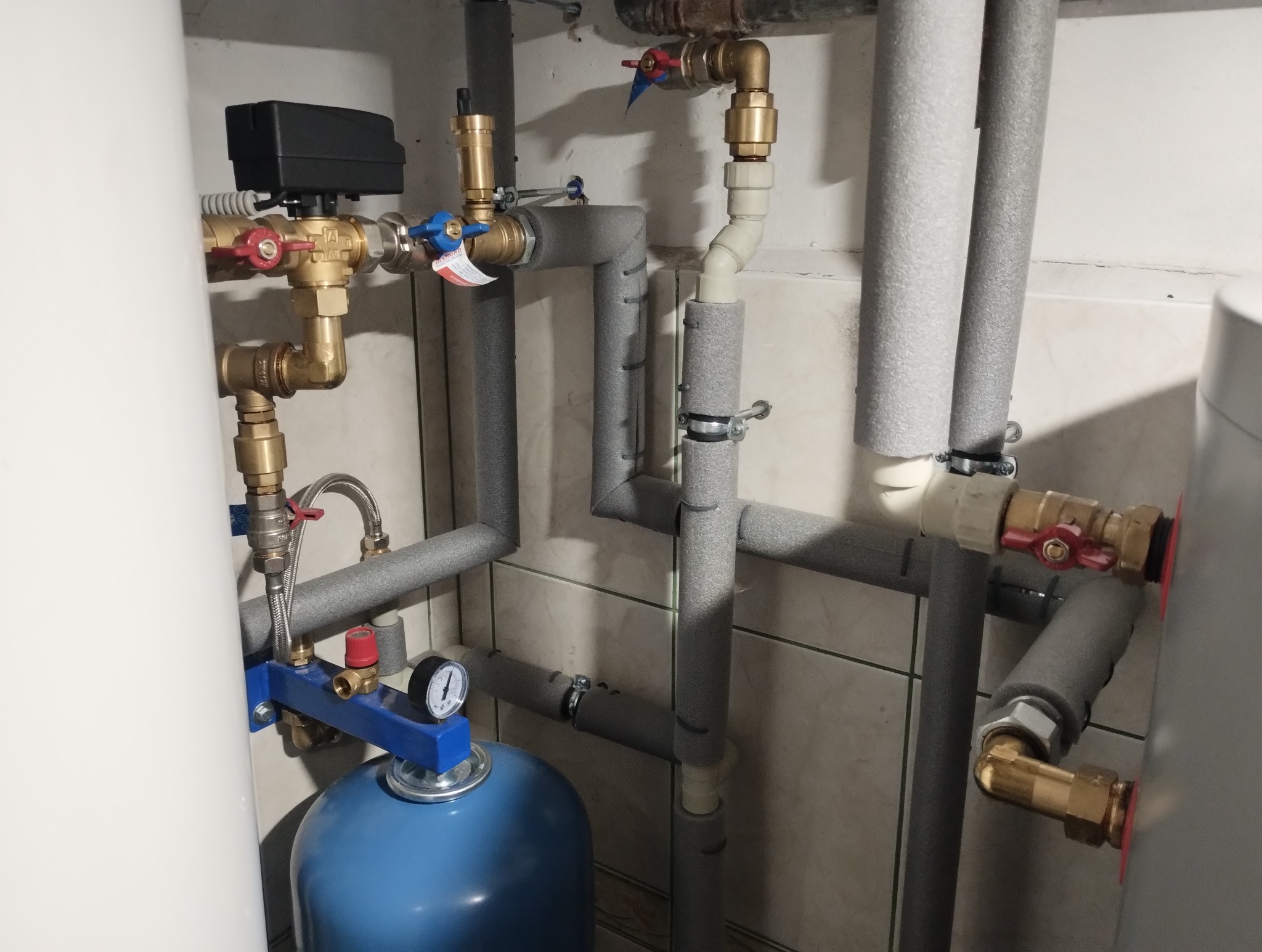 Instalacja grzewcza: rury w izolacji, zawory, manometr, niebieski zbiornik wyrównawczy, biały zasobnik ciepłej wody, płytki ceramiczne na ścianie.