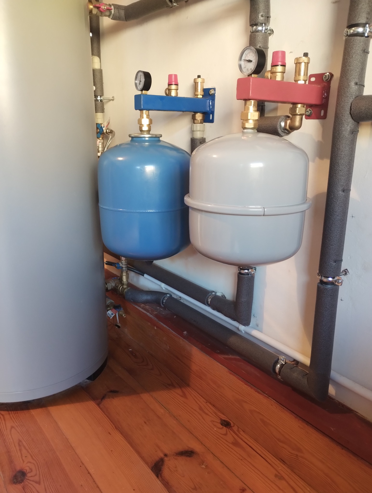 Dwa naczynia wzbiorcze, niebieskie i szare, zamontowane na ścianie z rurami w izolacji, na tle drewnianej podłogi.