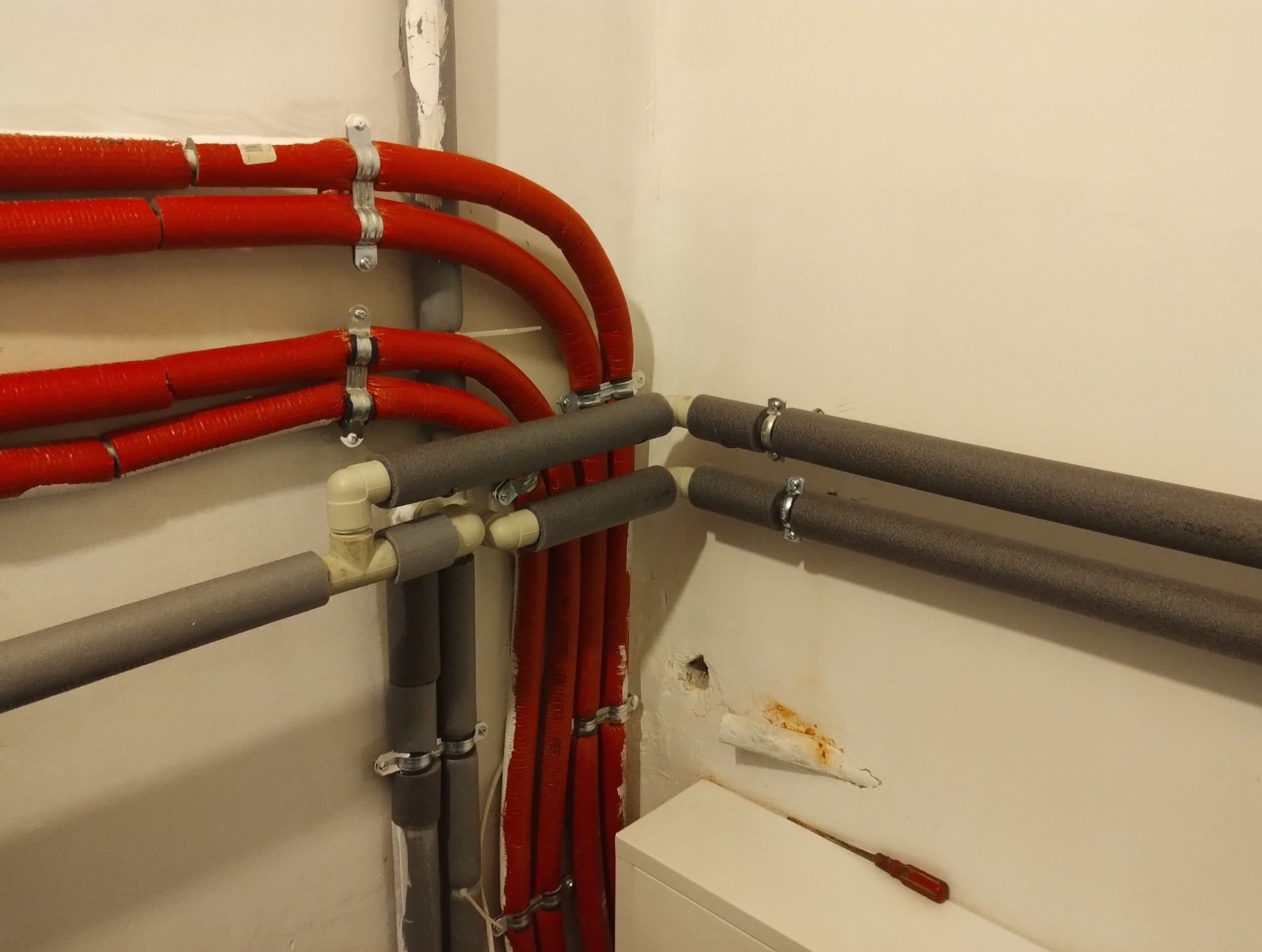 Rury grzewcze w izolacji, czerwone i szare, zamocowane do ściany za pomocą metalowych uchwytów, widoczny fragment grzejnika i śrubokręt z czerwoną rączką.