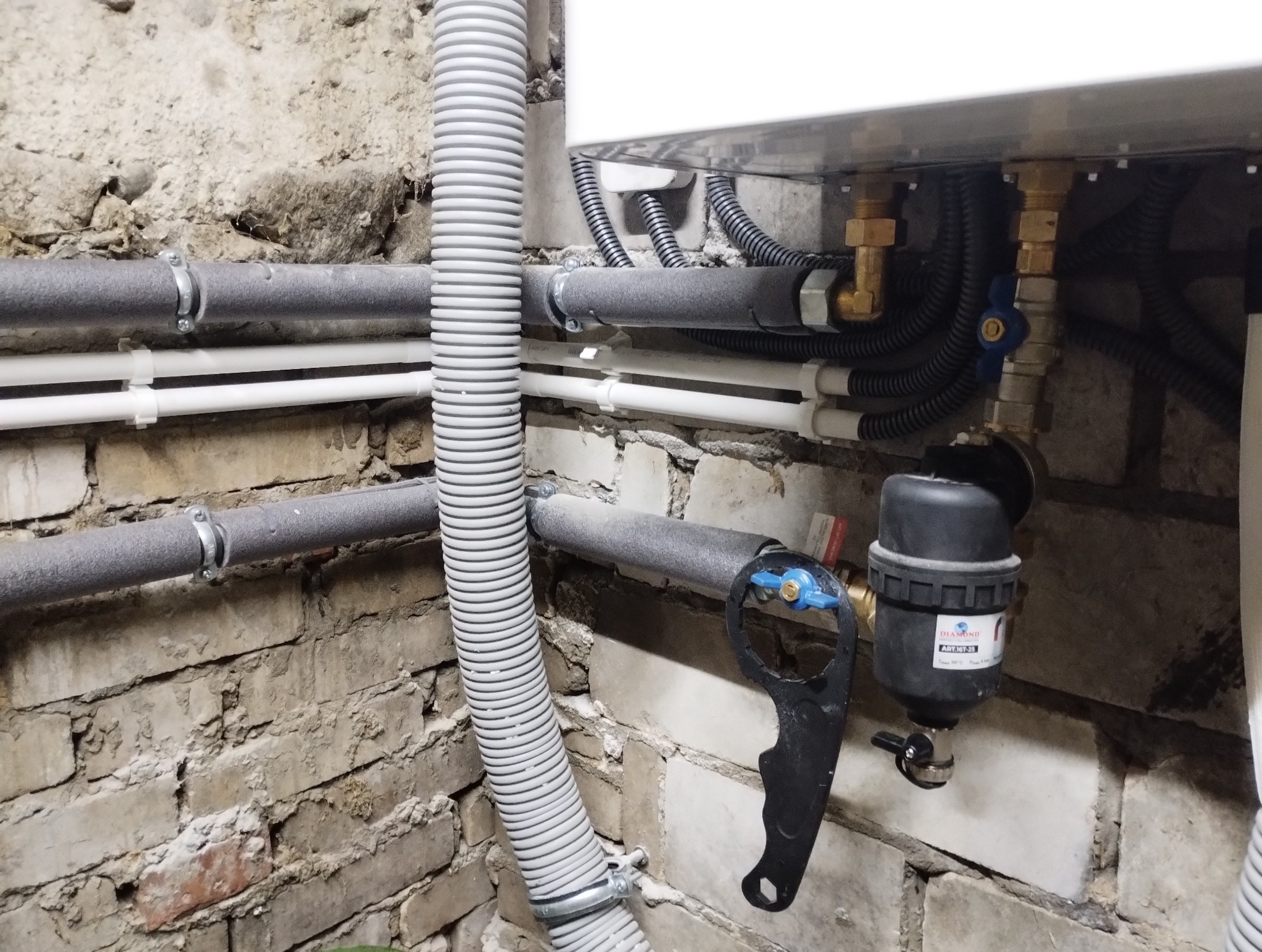 Fragment instalacji grzewczej z rurami izolowanymi termicznie, filtrem mechanicznym i kluczem do zaworów na tle ceglanej ściany.