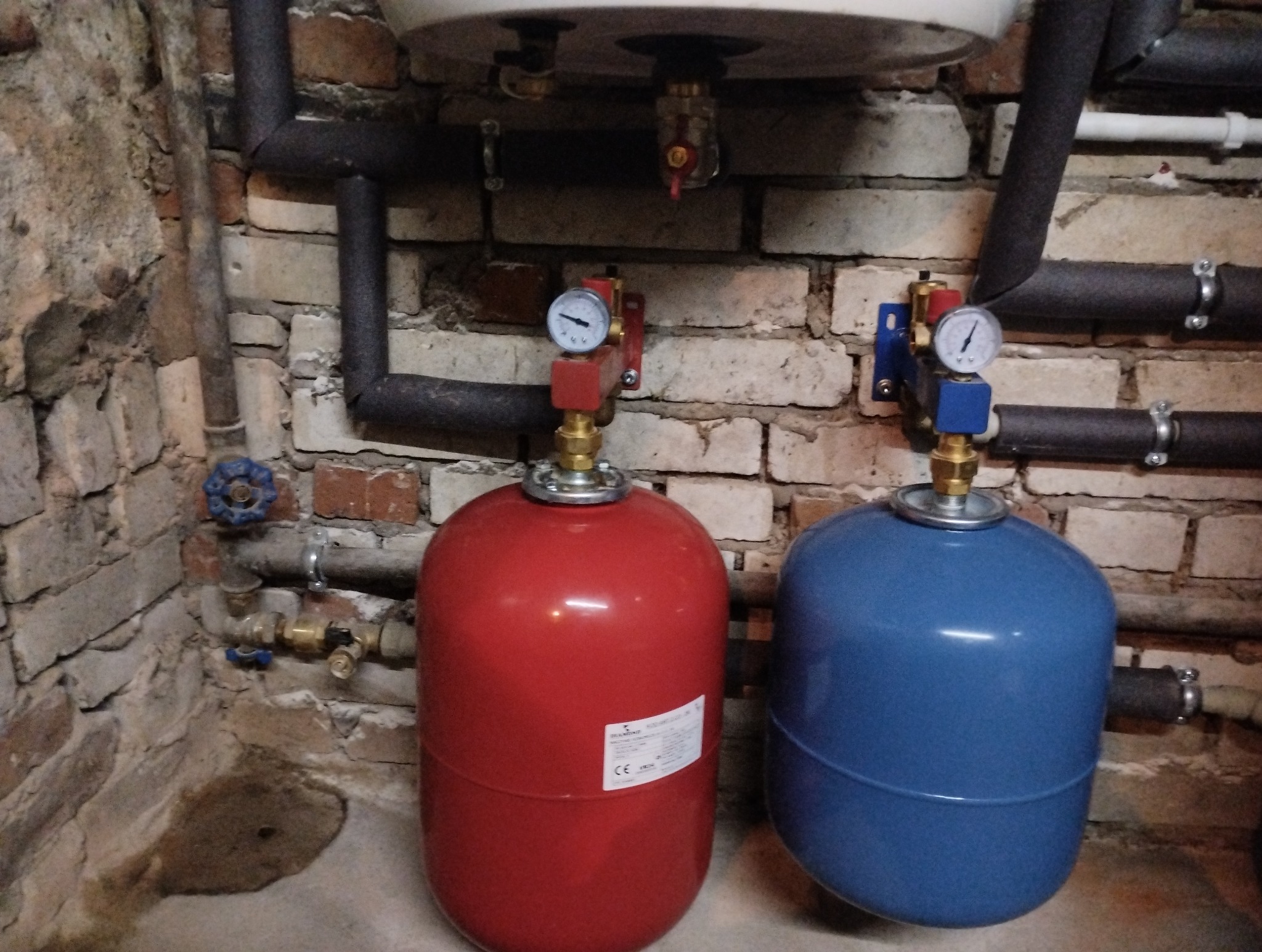 Czerwony i niebieski zbiornik wyrównawczy z manometrami w kotłowni z instalacją rurową na tle ceglanej ściany.