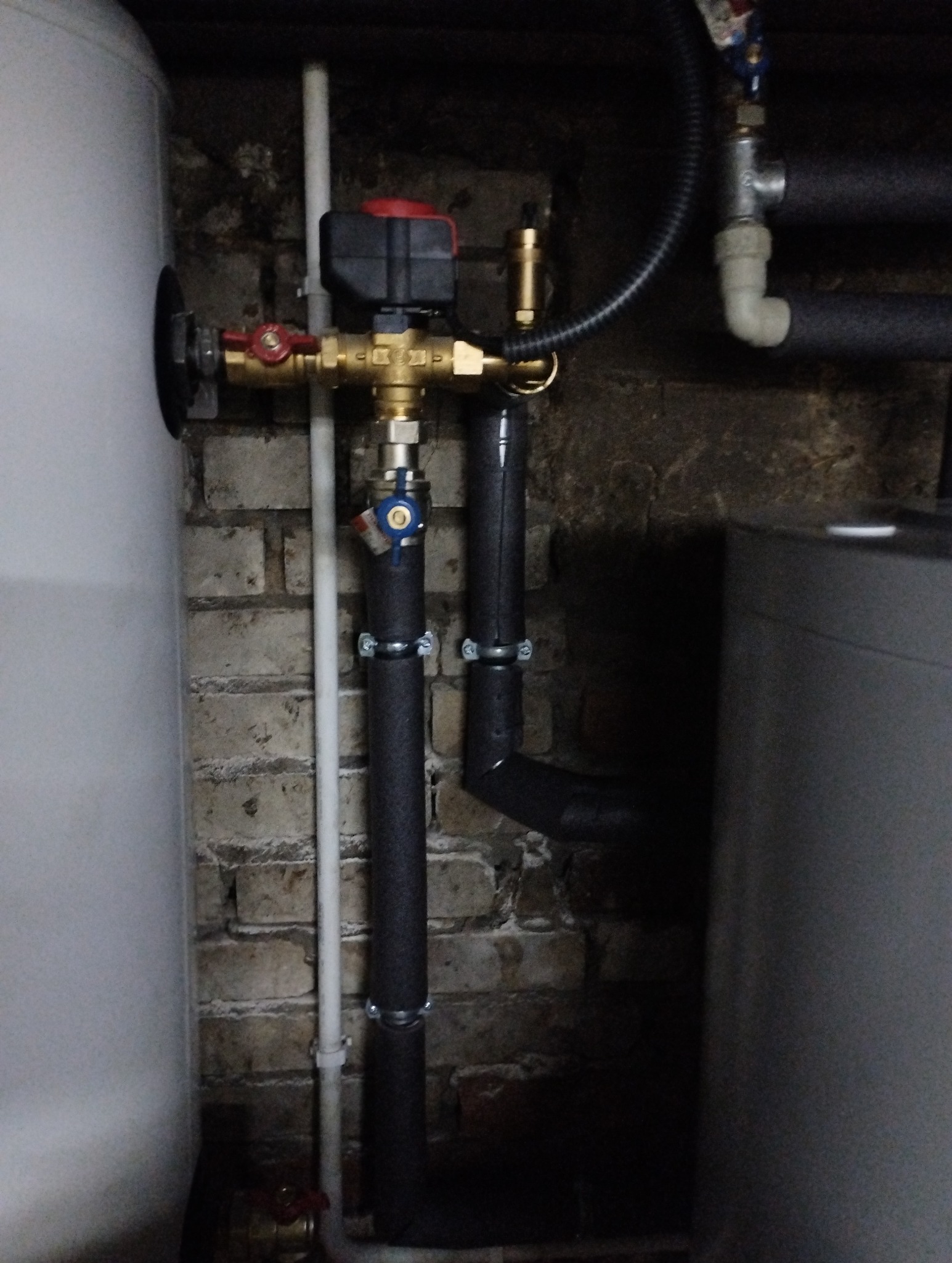 Fragment instalacji grzewczej z widocznymi rurami, zaworami i zbiornikiem akumulacyjnym na tle ceglanej ściany.