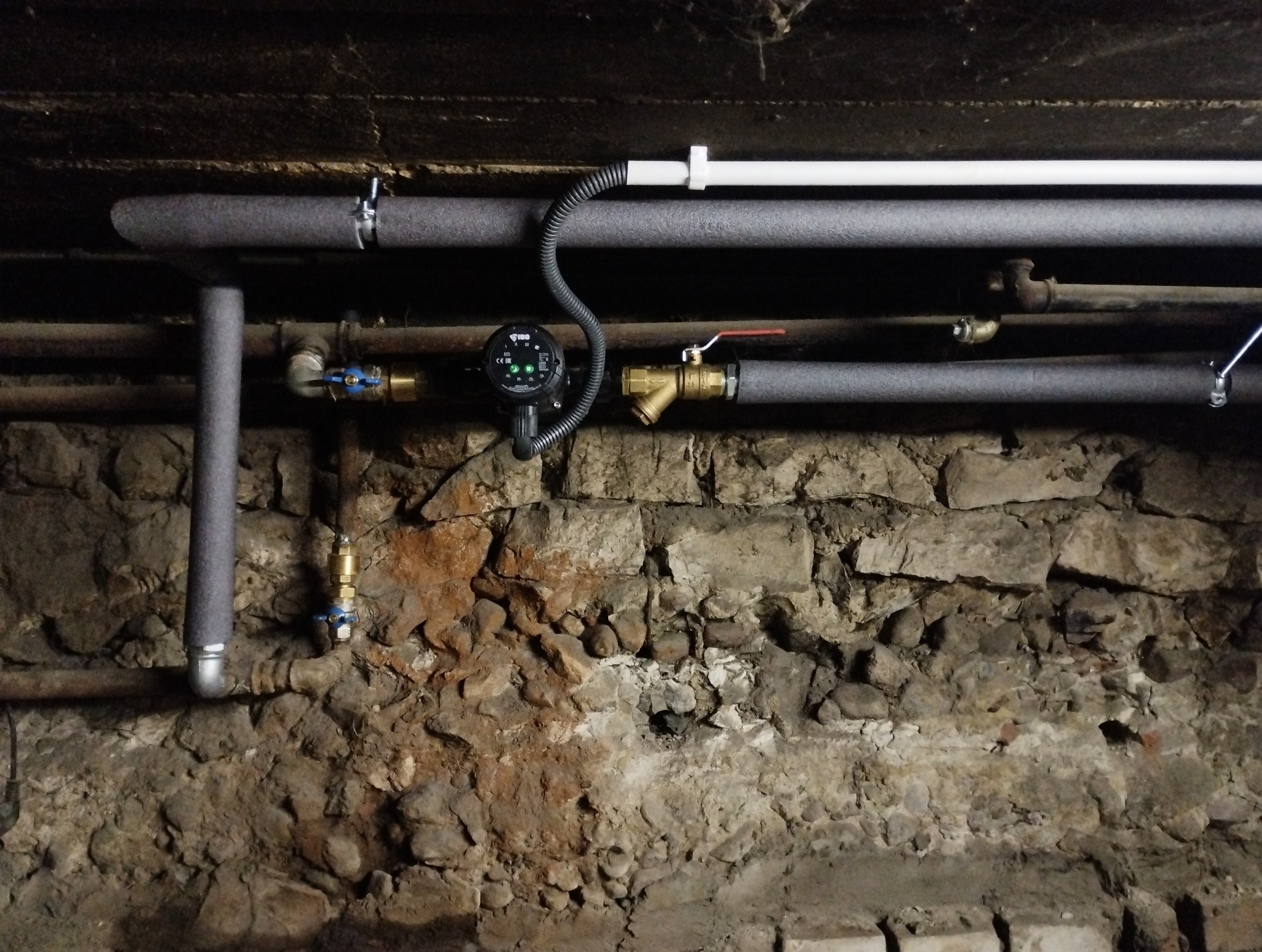 Instalacja rur grzewczych z izolacją, zaworami i pompą obiegową na tle kamiennej ściany w piwnicy.